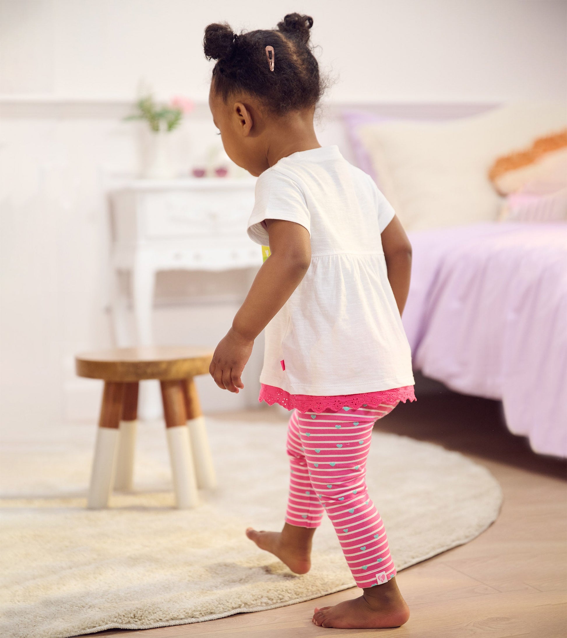 White t-shirt with rainbow eyelet trim paired with pink striped leggings featuring small hearts, worn by a toddler.