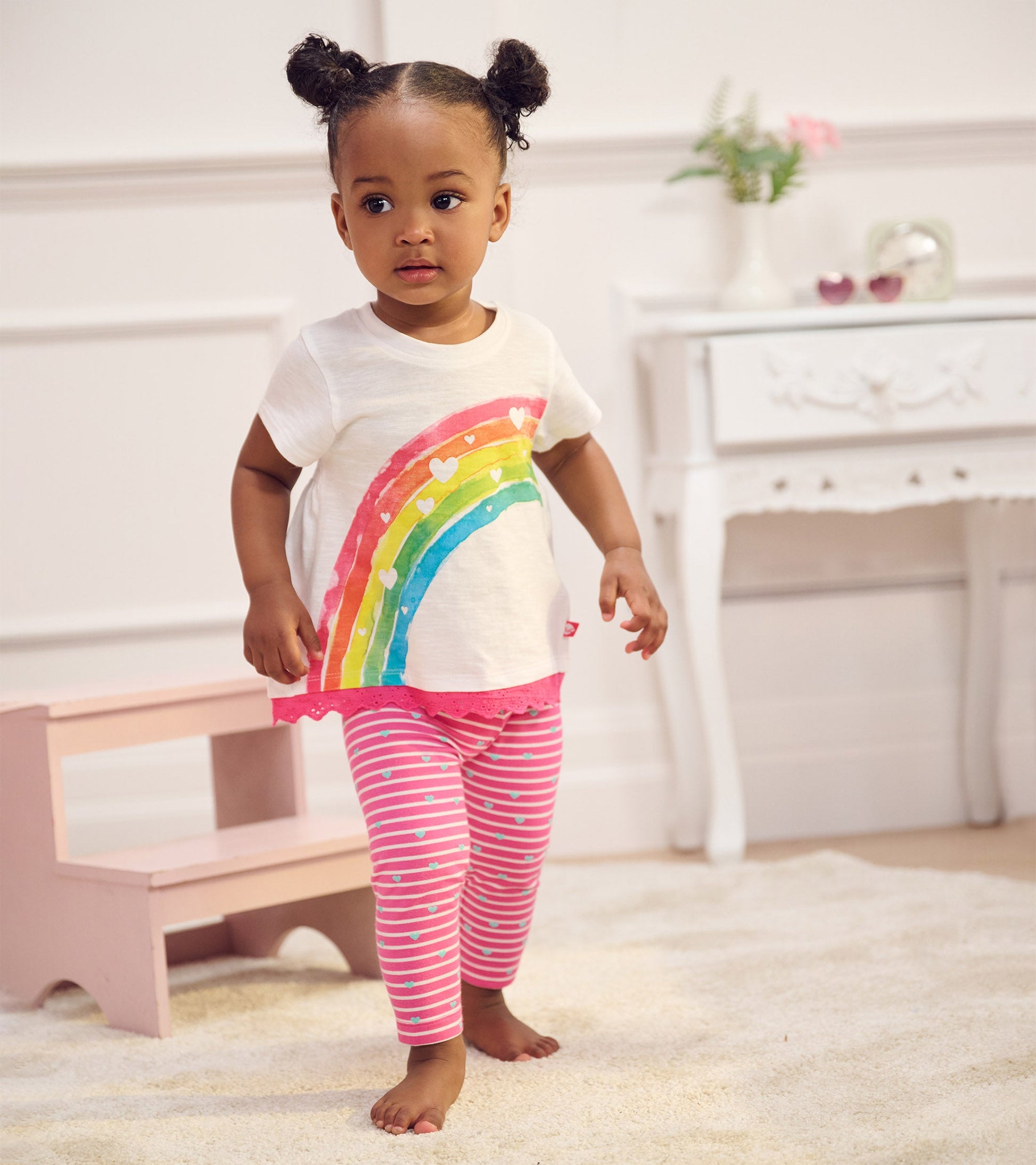 White t-shirt with rainbow and heart design, paired with pink striped leggings, worn by a toddler girl.