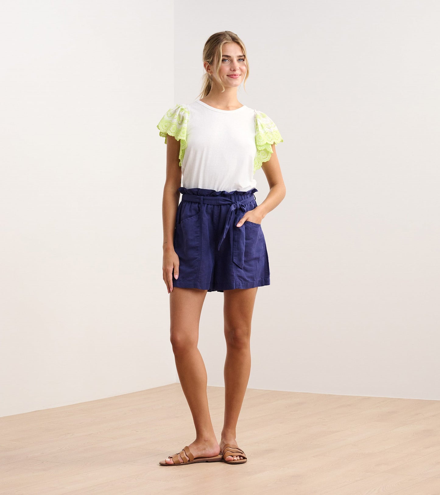 White tank top with eyelet sleeves, paired with navy shorts, worn by a model standing on a light wooden floor.