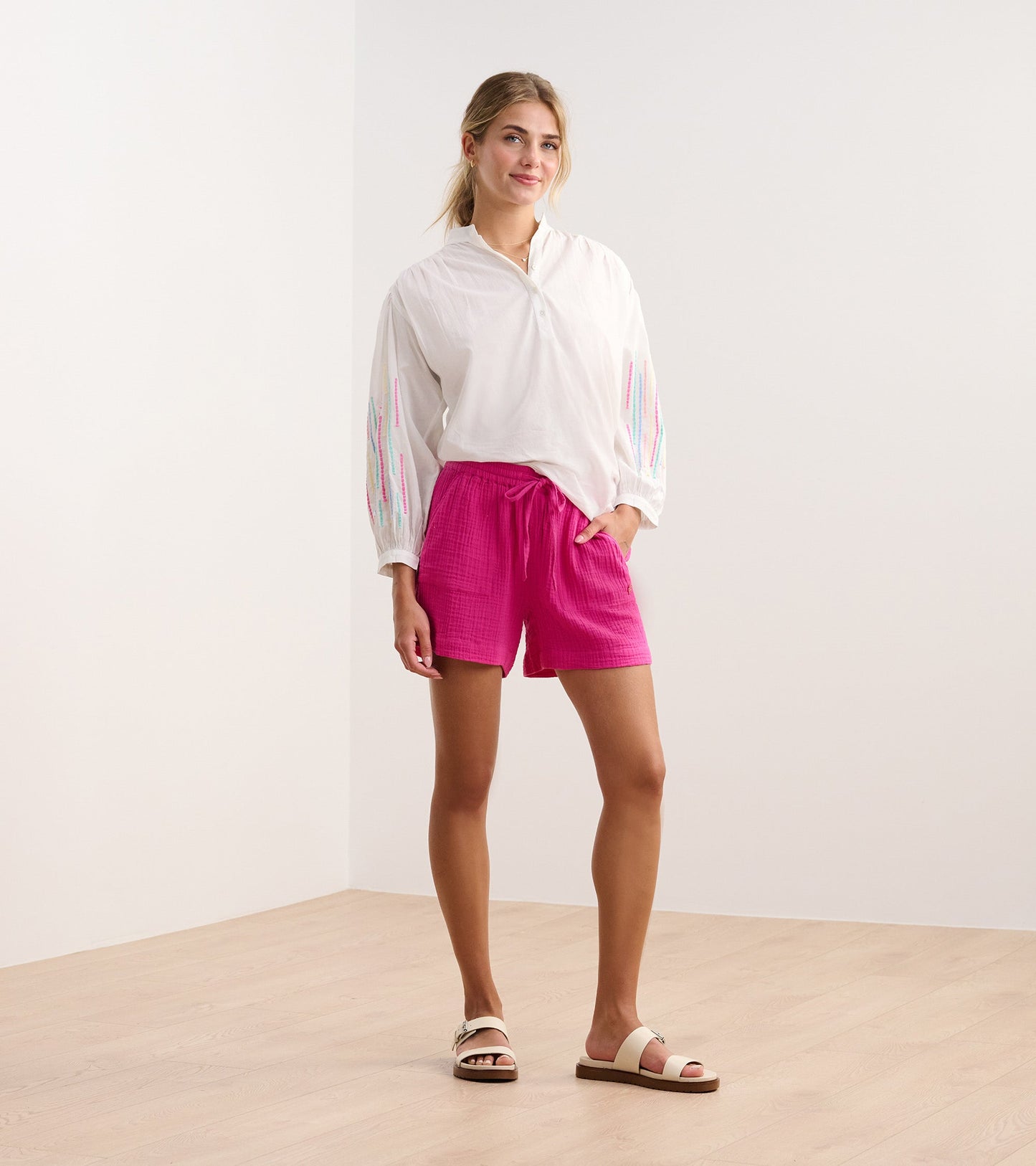 White gauze tunic with colorful embroidery on sleeves, paired with pink shorts and sandals, standing on a wooden floor.