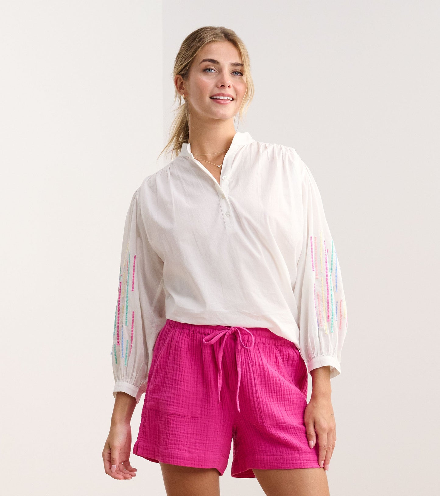 White gauze tunic with colorful embroidery on sleeves, paired with pink shorts, styled for a casual look.