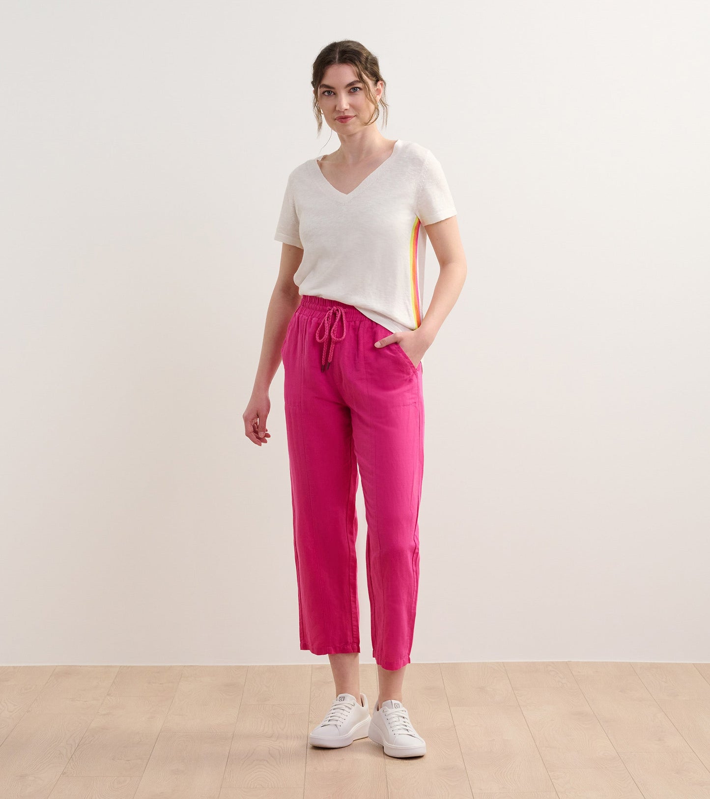 White V-neck knit tee with short sleeves, paired with pink drawstring pants, worn by a model standing on a wooden floor.