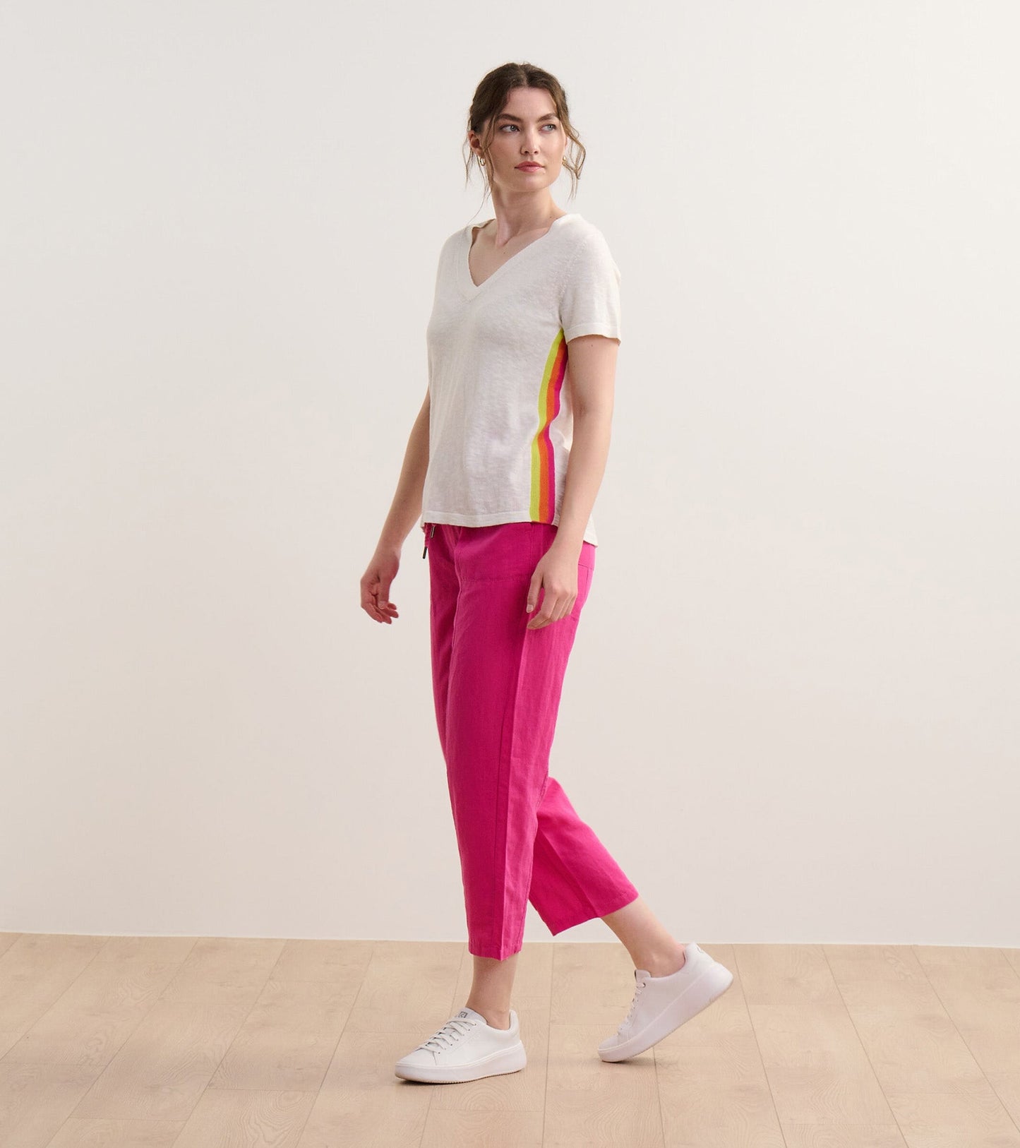 White V-neck knit tee with colorful side stripes, paired with pink pants, modeled by a woman standing on a light wooden floo…