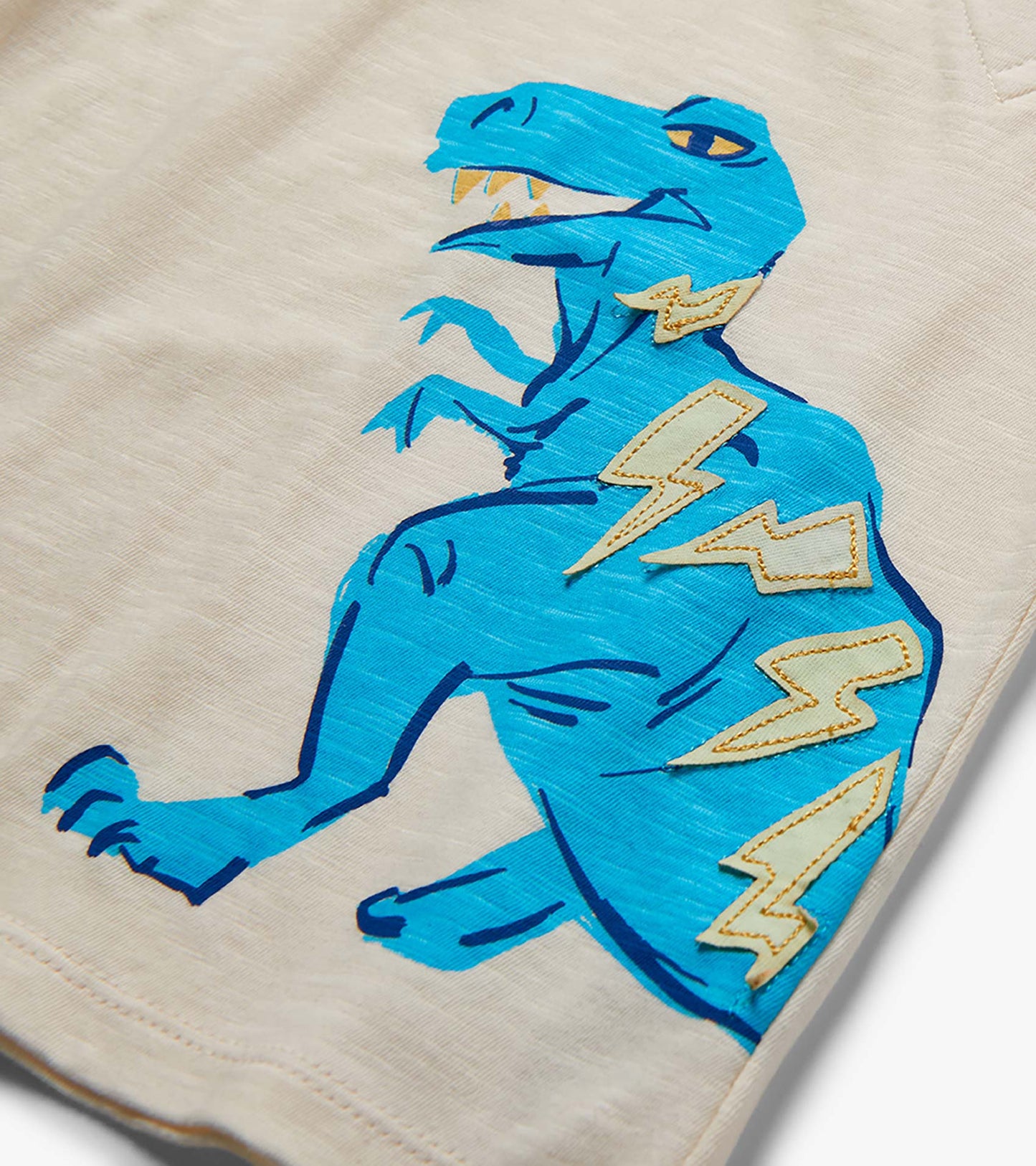 Close-up of beige shorts with a blue dinosaur graphic and lightning bolt patches