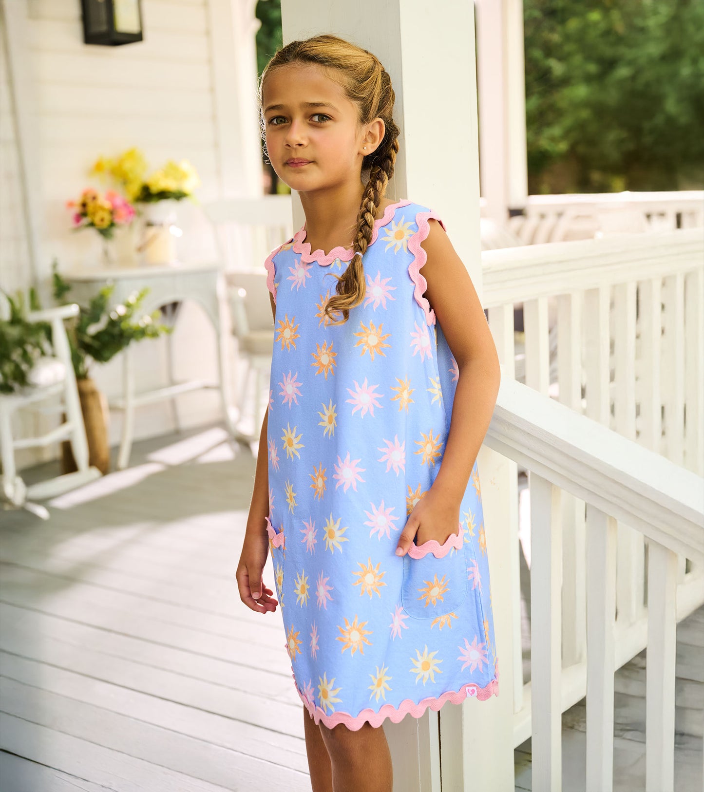 Side view of blue pinafore dress with faded sun print, pink rickrack trim, and front patch pocket
