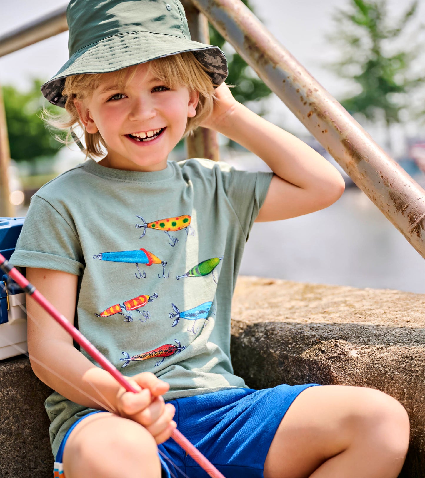 Close-up of sage green boys tee with multicolored fishing lure print, worn with a bucket hat