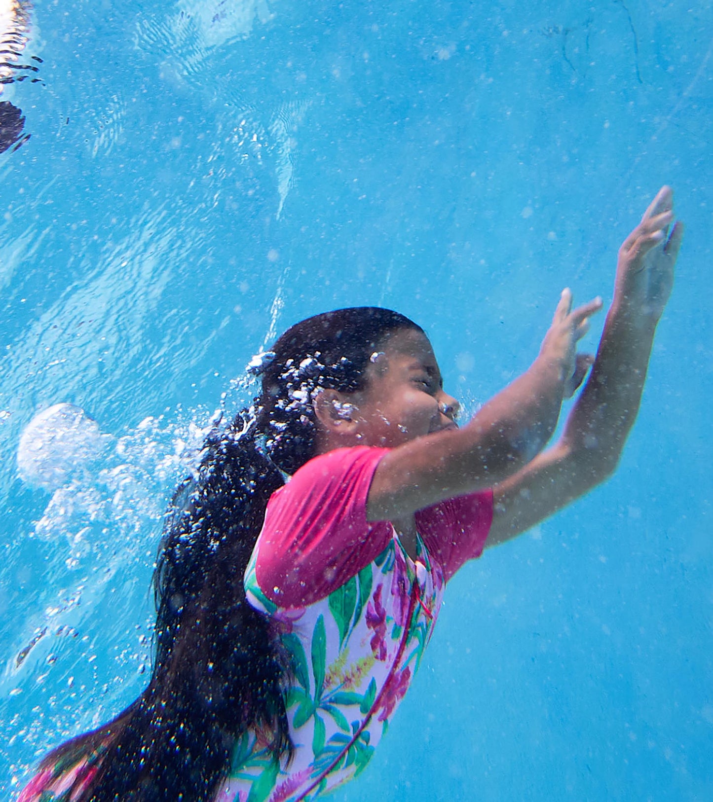 Girl wearing pink short-sleeve one-piece rashguard swimsuit with tropical floral print, swimming underwater