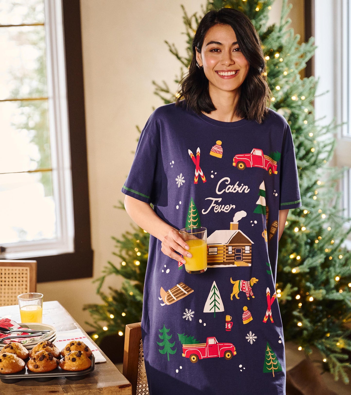Model wearing navy cabin print sleepshirt indoors near lit Christmas trees, holding a glass of orange juice