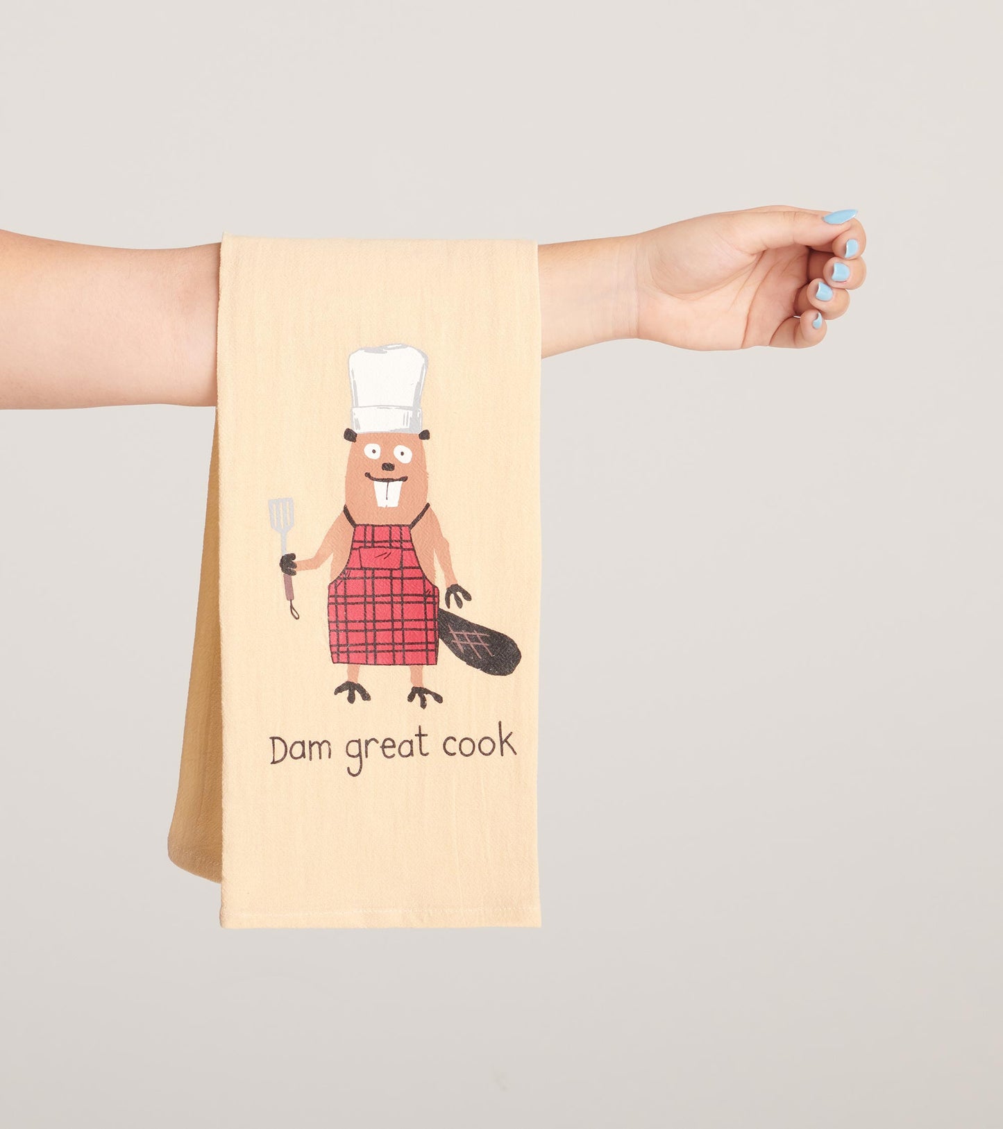 Beige tea towel featuring a cartoon beaver in a red plaid apron, holding a spatula with "Dam great cook" text.