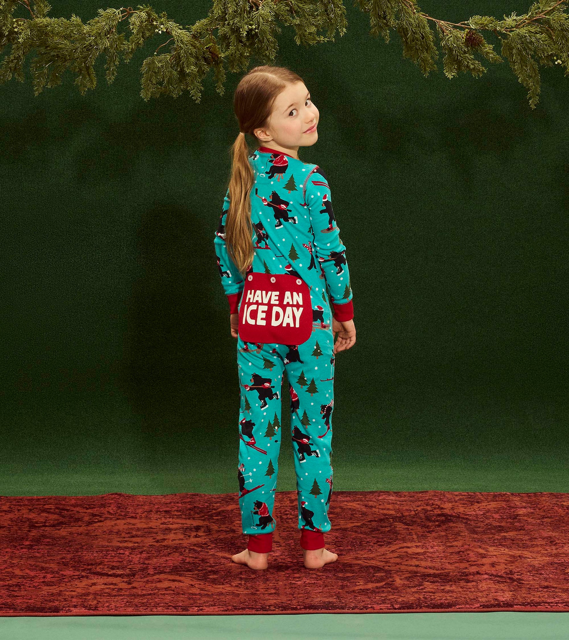 Turquoise winter onesie with playful patterns of trees and animals, featuring a red pocket with "HAVE AN ICE DAY" text.
