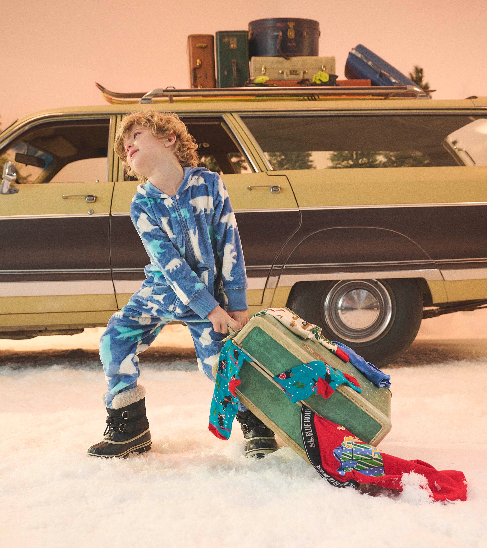 Blue fleece jumpsuit featuring polar bear patterns, worn by a child in snow, with a suitcase and vintage car in the backgrou…