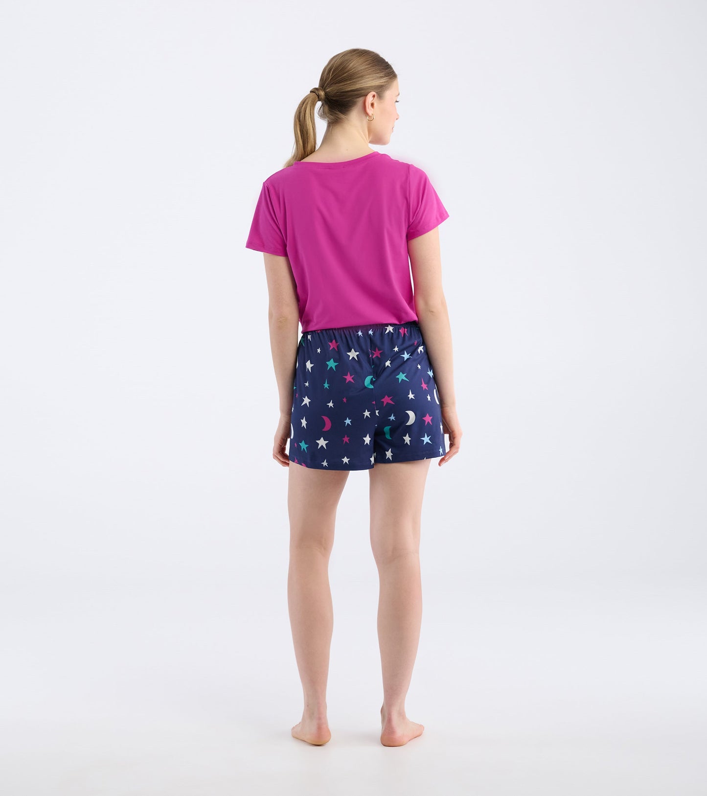 Back view of a woman wearing pink top and navy pajama shorts with colorful star and moon pattern.