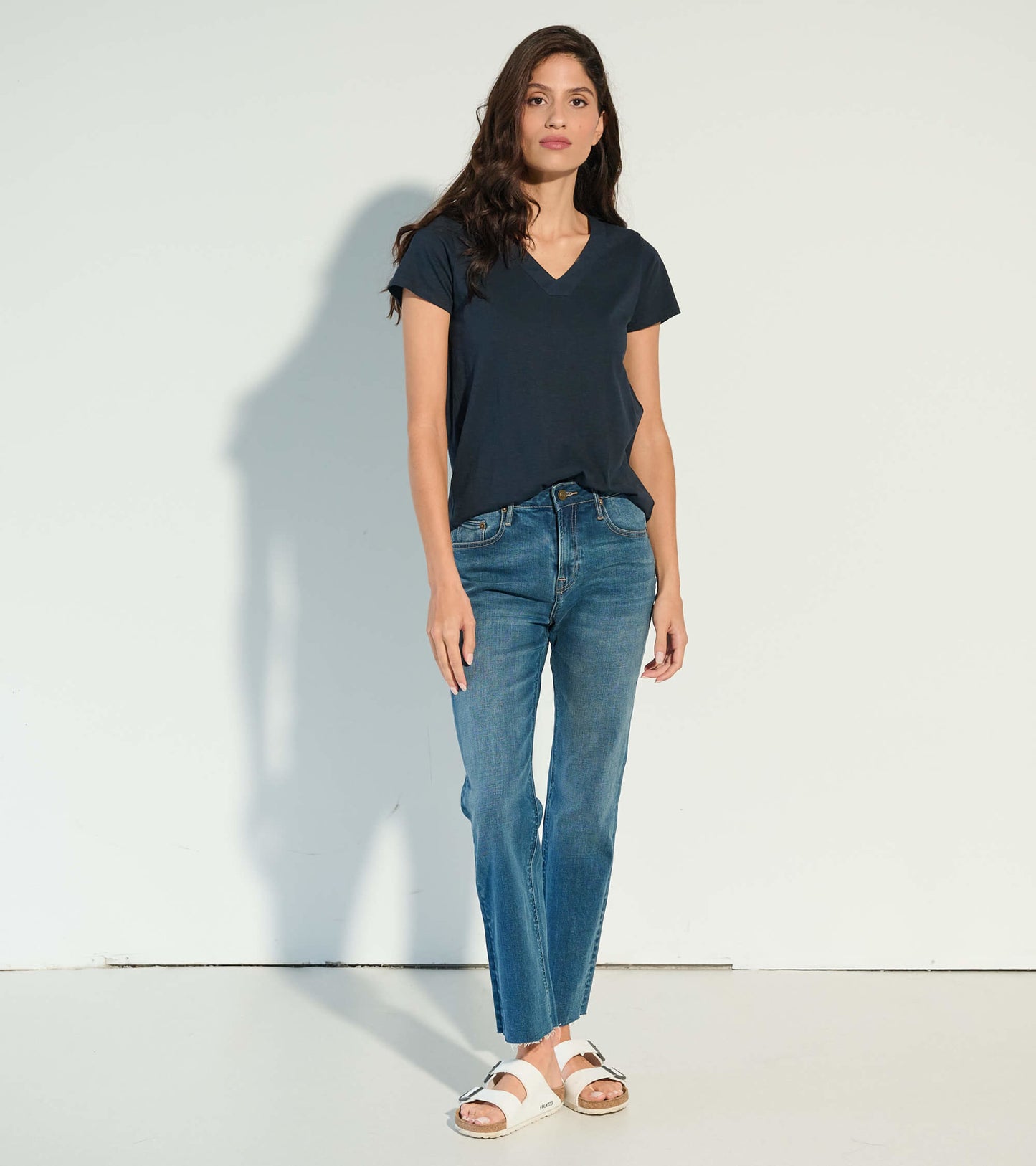 Black v-neck tee with short sleeves, paired with blue jeans and white sandals, standing against a light background.