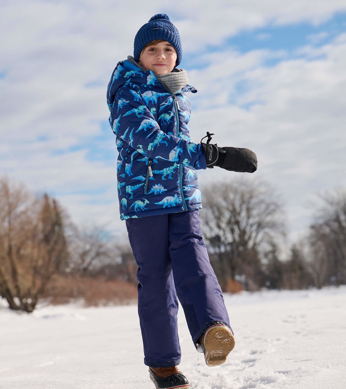 Blue puffer jacket with dinosaur print, worn by a child in snow, paired with dark pants, gloves, and a knit hat.