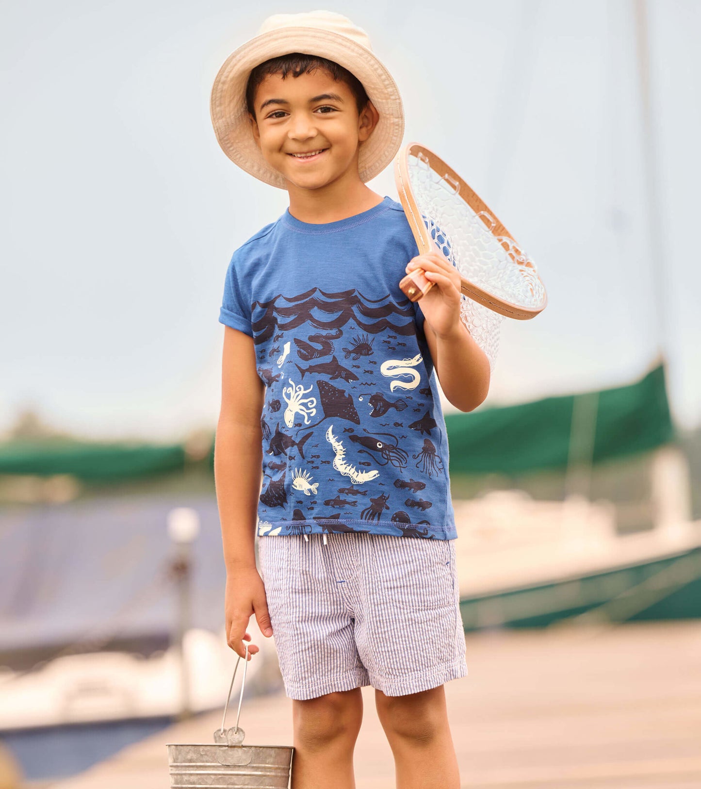 Blue graphic tee featuring ocean-themed designs, worn by a child holding a fishing net and bucket, paired with striped short…