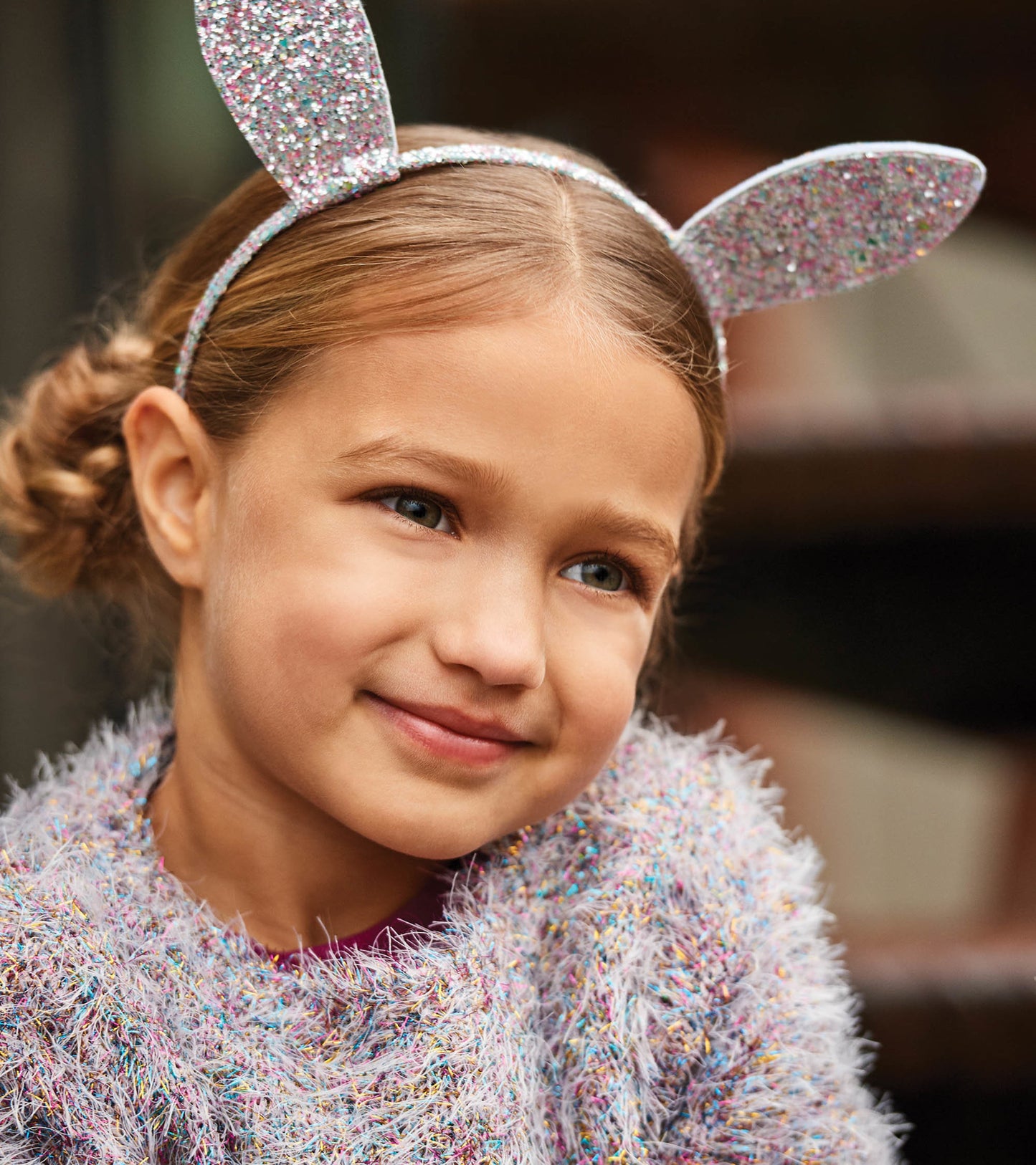 Glittery pink and silver bunny ears headband worn by a girl with a fluffy, colorful sweater.