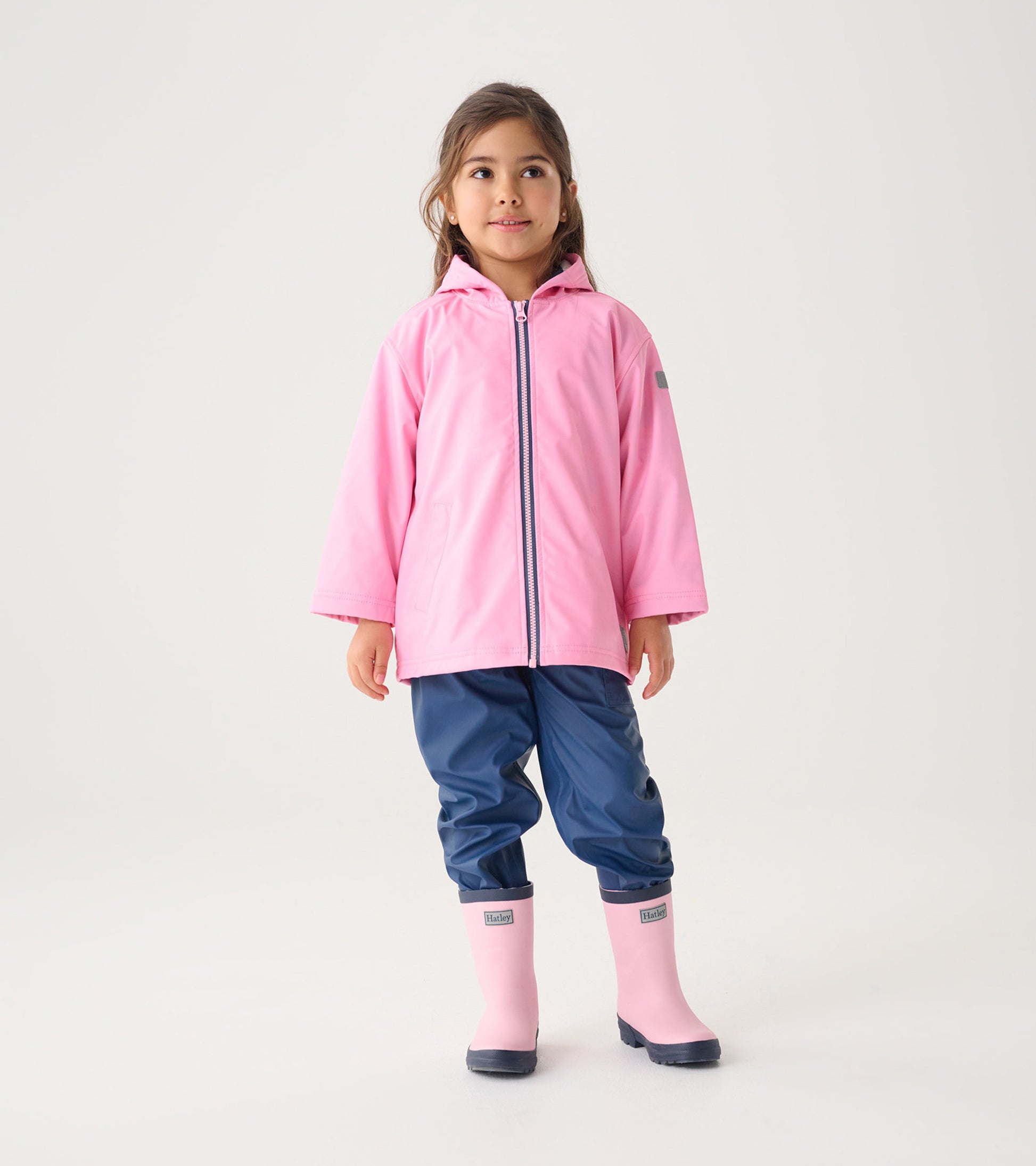 Pink zip-up rain jacket with a hood, worn by a girl, paired with navy pants and pink rain boots.