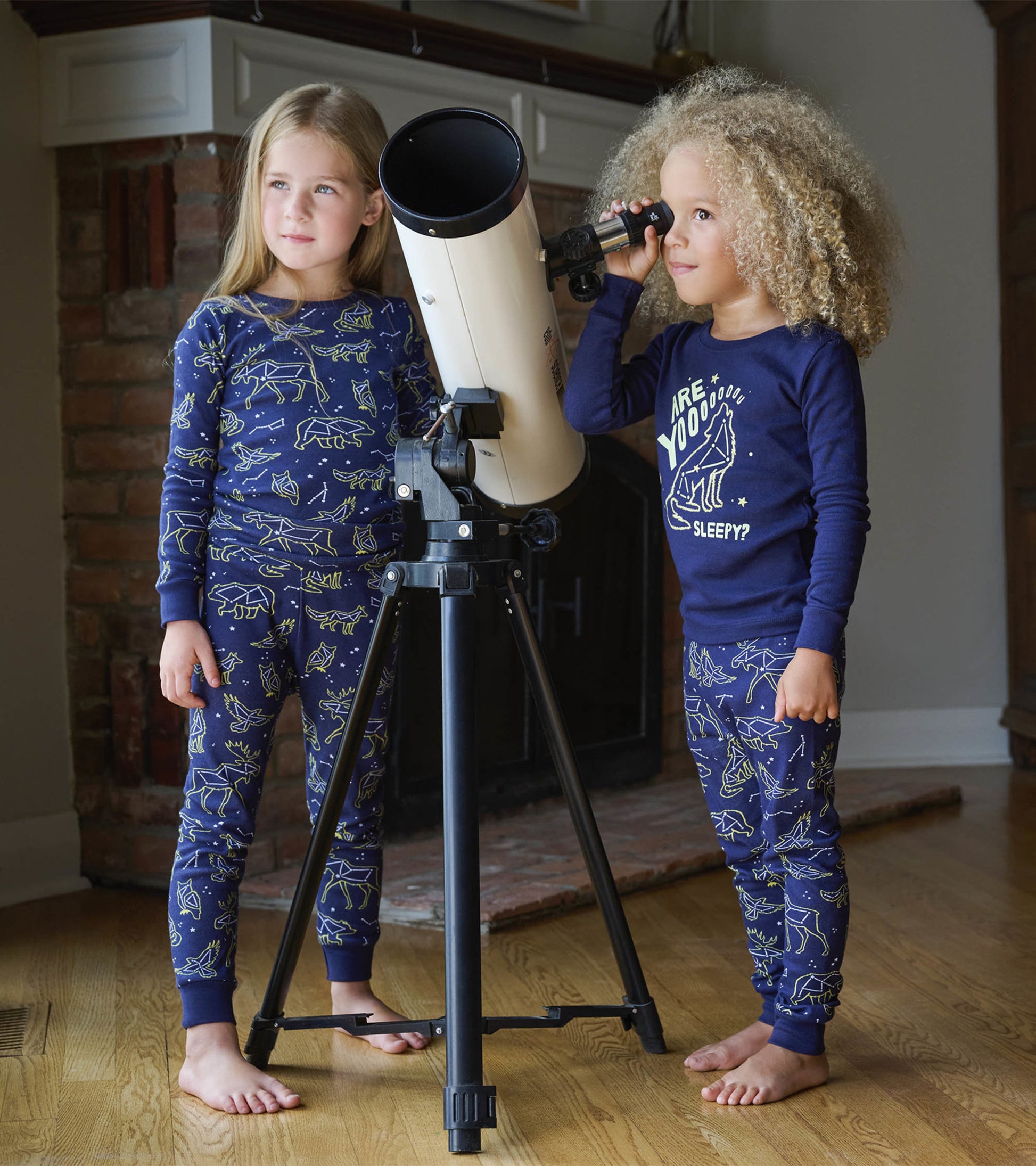 Navy blue kids' pajama set with constellation patterns, featuring long sleeves and pants, worn by two children near a telesc…