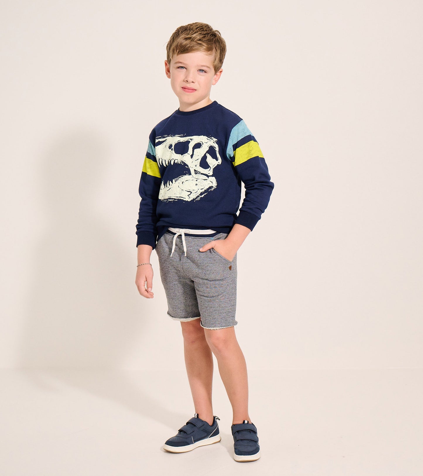 Navy pullover sweatshirt featuring a dinosaur skull graphic and colorful striped sleeves, worn with gray shorts.