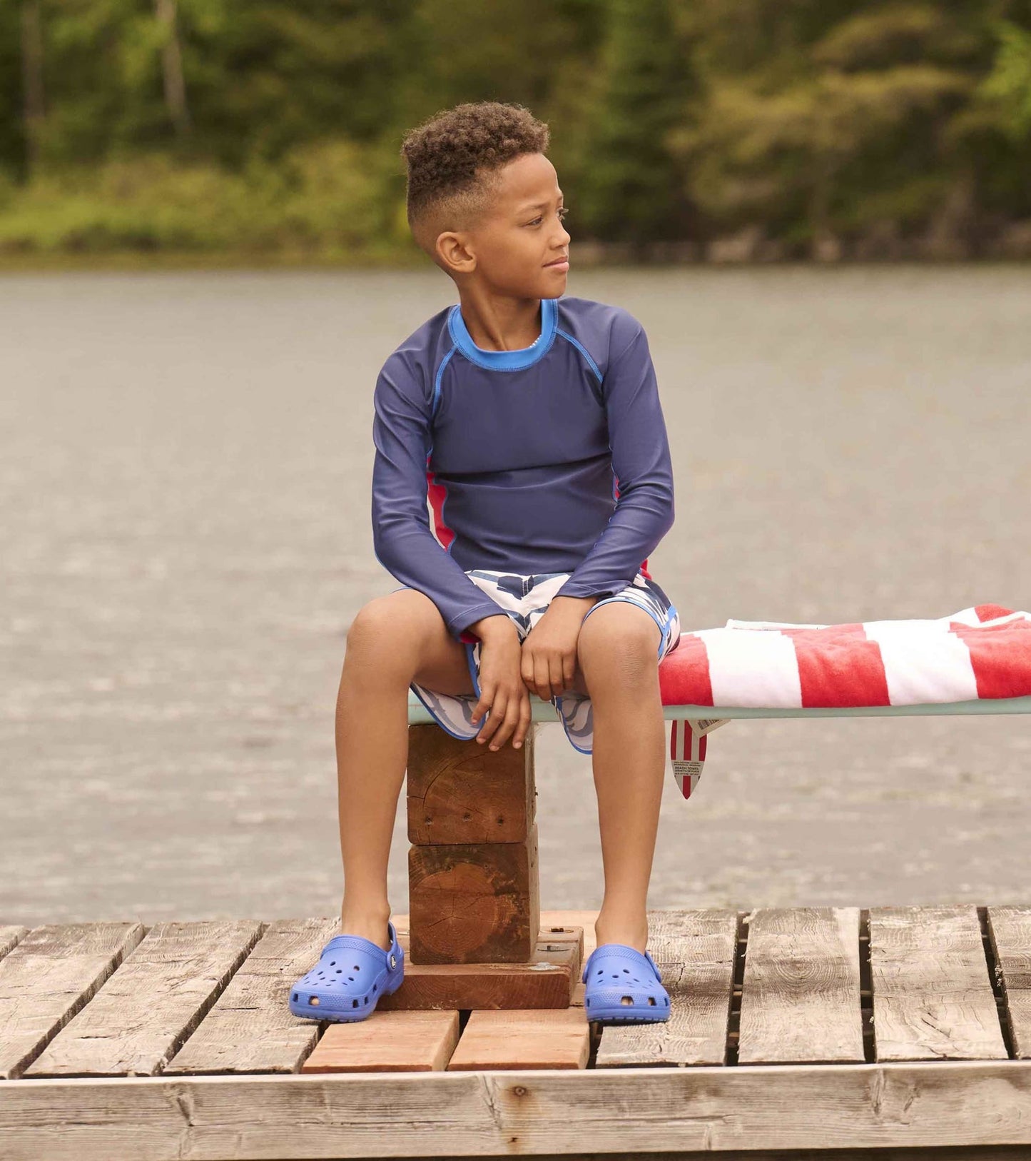Nautical long sleeve rashguard in navy with red accents, worn by a boy sitting on a dock, paired with blue crocs.