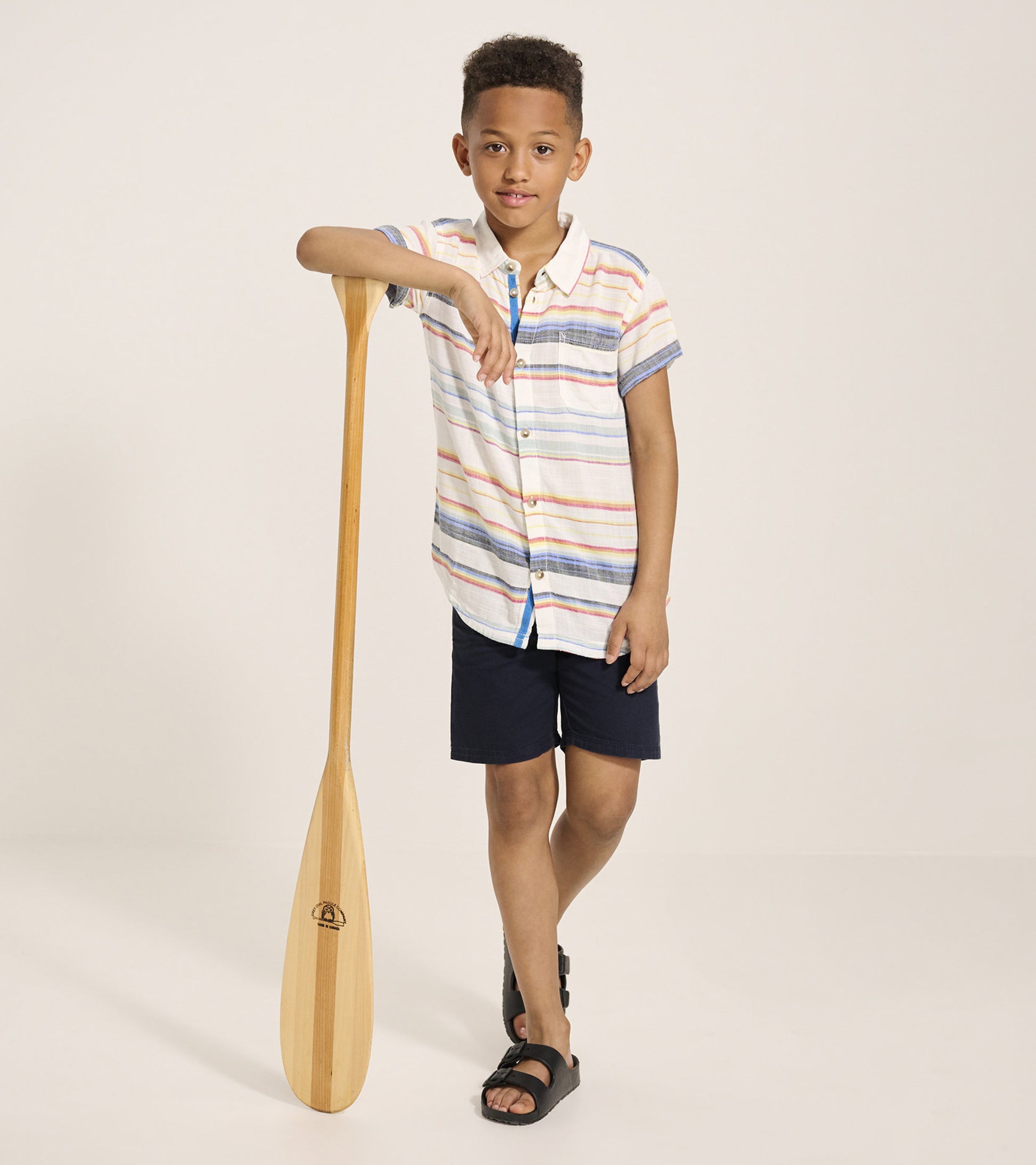 Short sleeve button-down shirt with colorful stripes, paired with navy shorts, worn by a boy holding a wooden paddle.