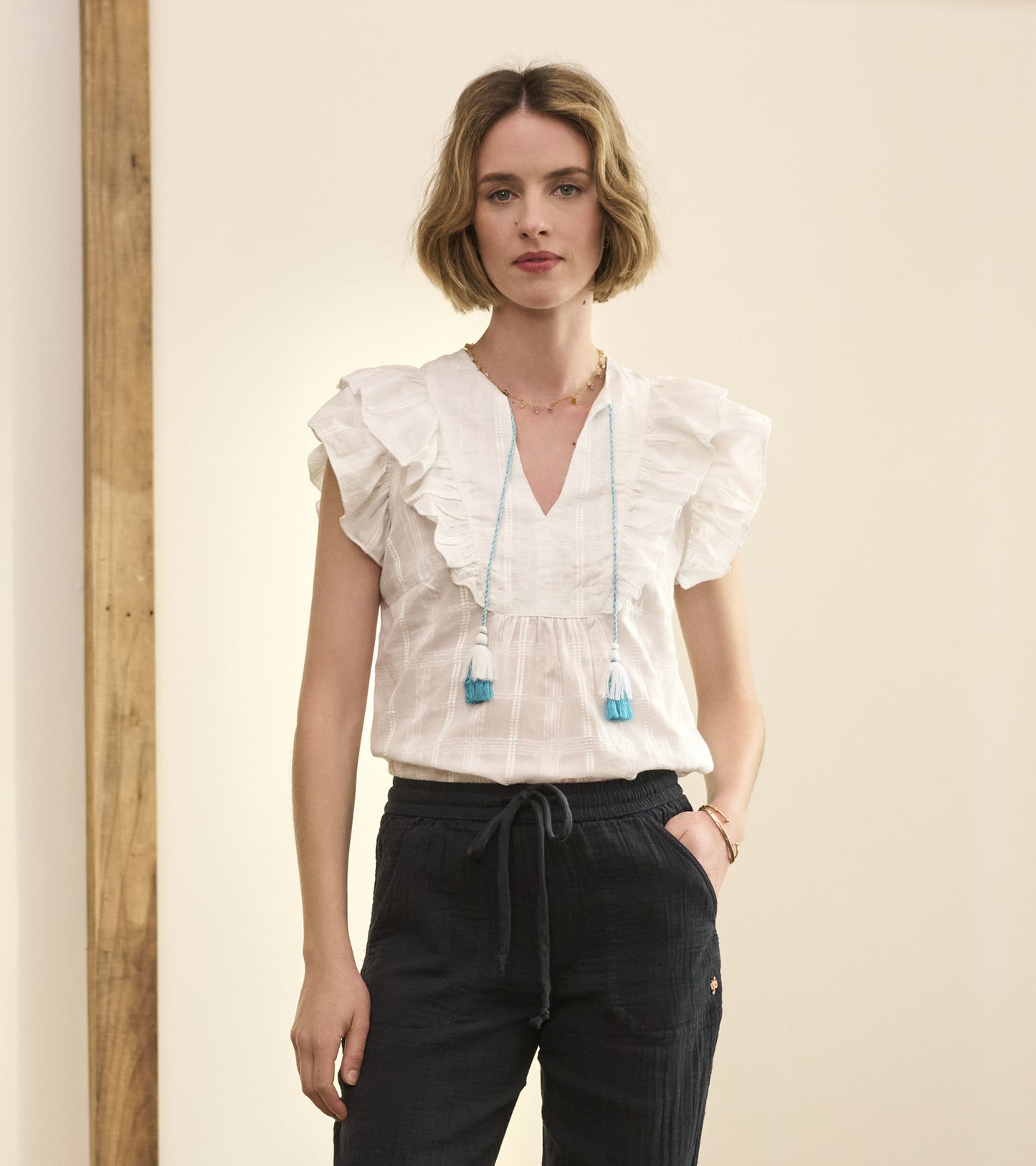 White blouse with ruffled sleeves and a V-neck, paired with dark drawstring pants. Model stands against a neutral background.