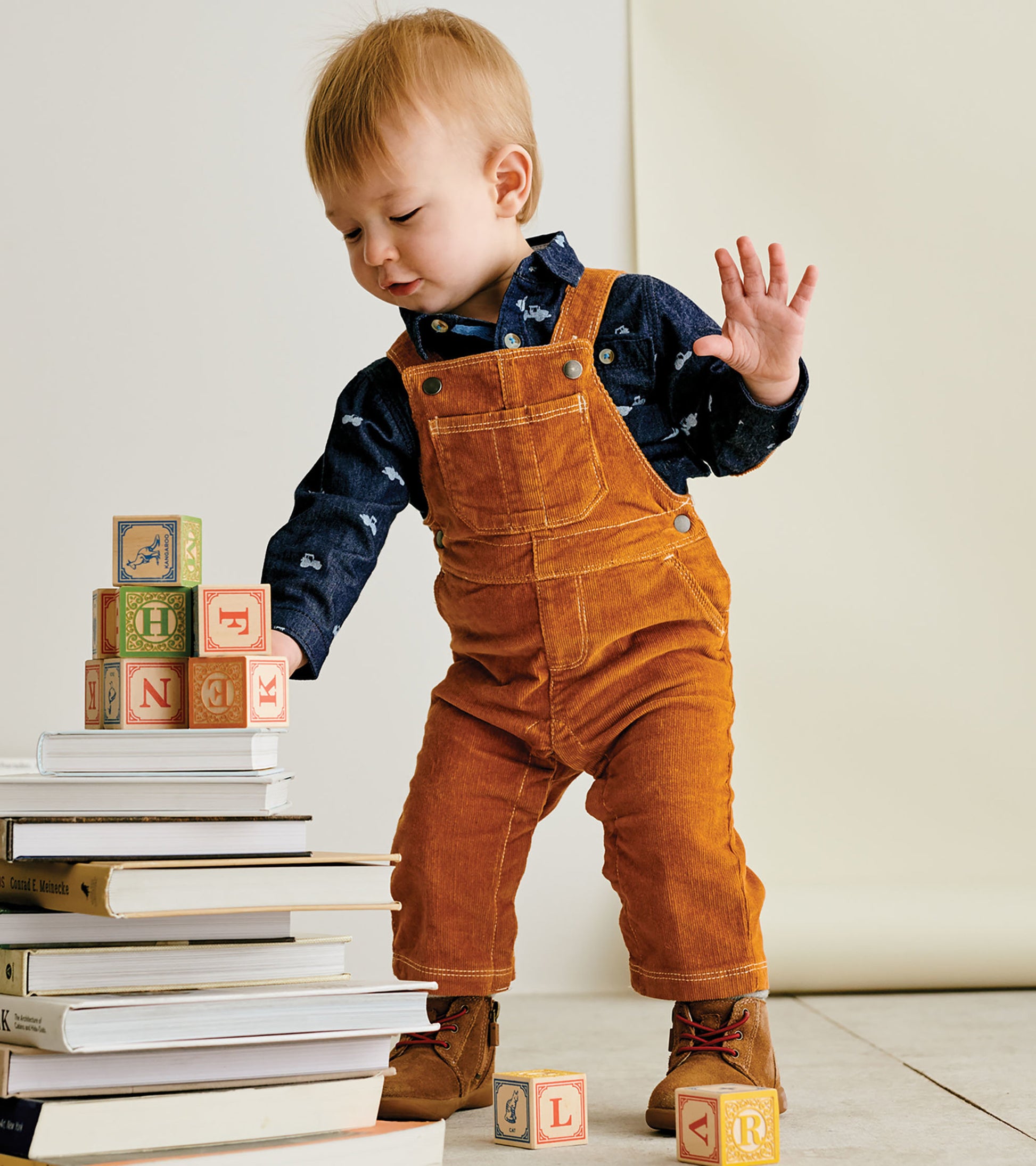 Pecan brown corduroy overalls on a toddler, paired with a navy shirt, playing with building blocks and books.