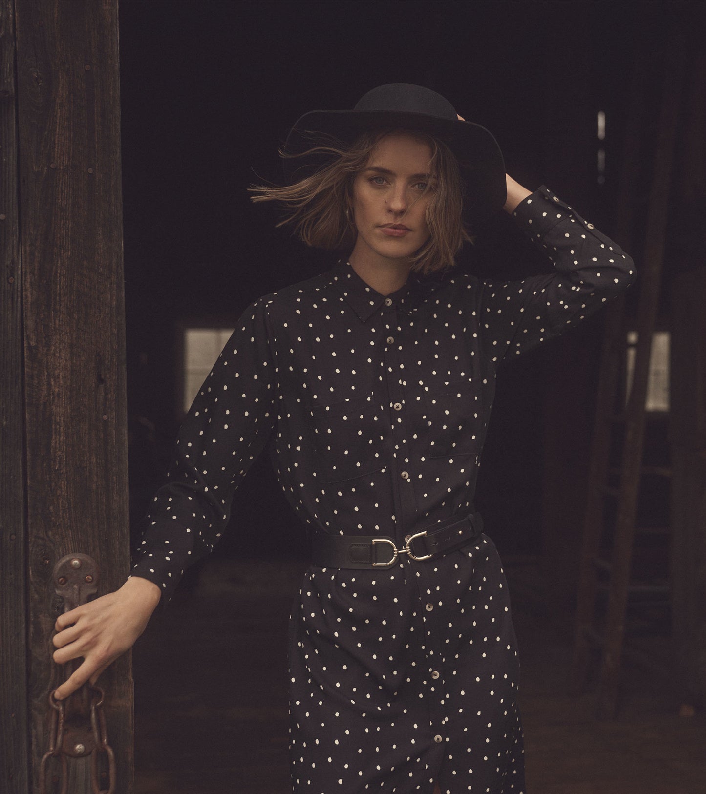 Black midi shirt dress with white polka dots, button-up front, and belted waist, styled with a wide-brimmed hat.