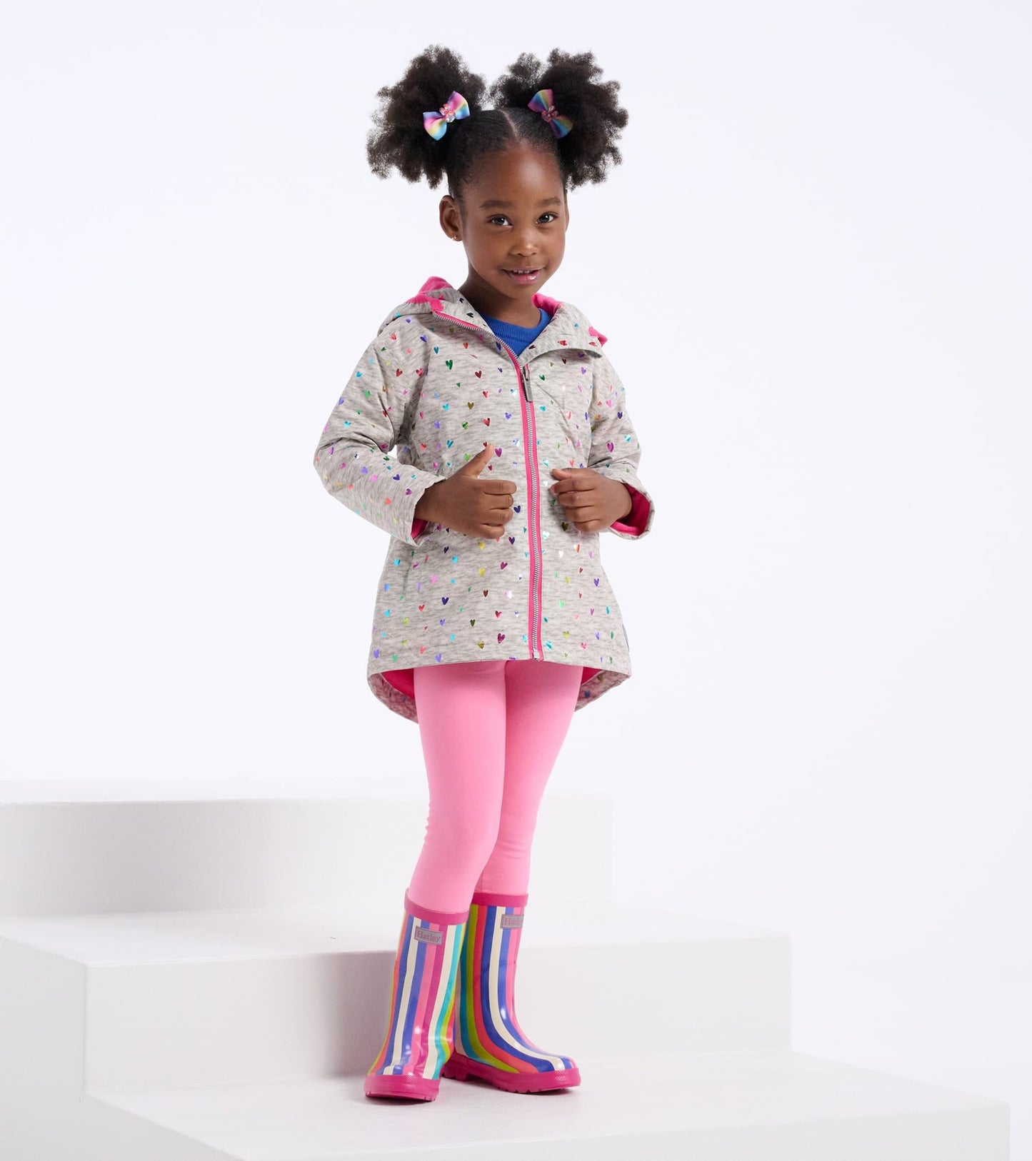 Lightweight zip-up rain jacket with colorful confetti hearts pattern, paired with pink leggings and striped rain boots.