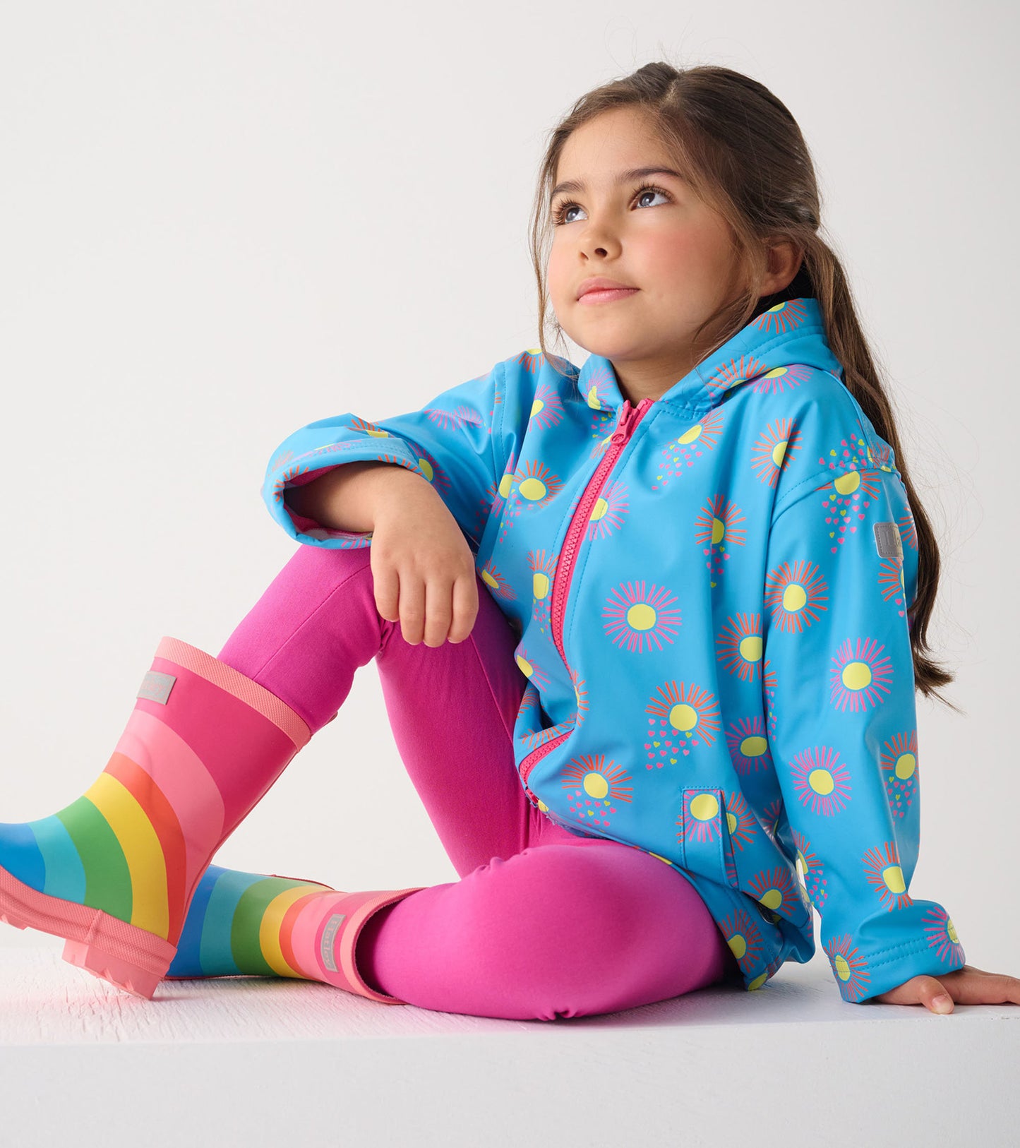 Blue zip-up rain jacket with sun patterns, paired with pink leggings and rainbow rain boots, worn by a seated girl.
