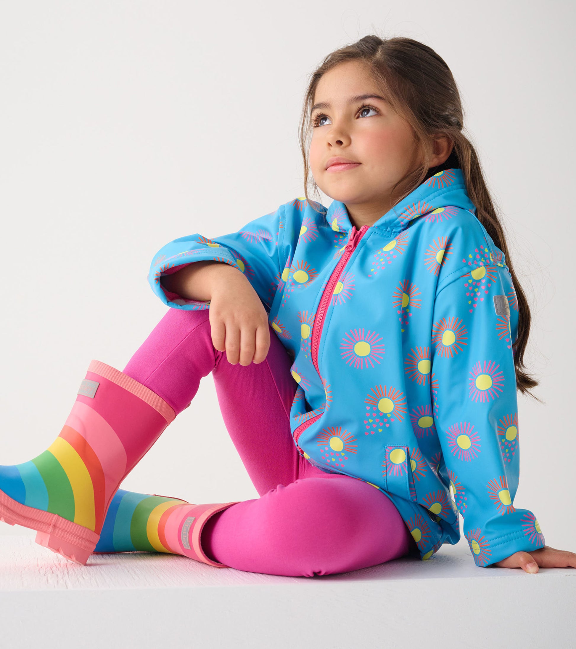 Blue zip-up rain jacket with sun patterns, paired with pink leggings and rainbow rain boots, worn by a seated girl.