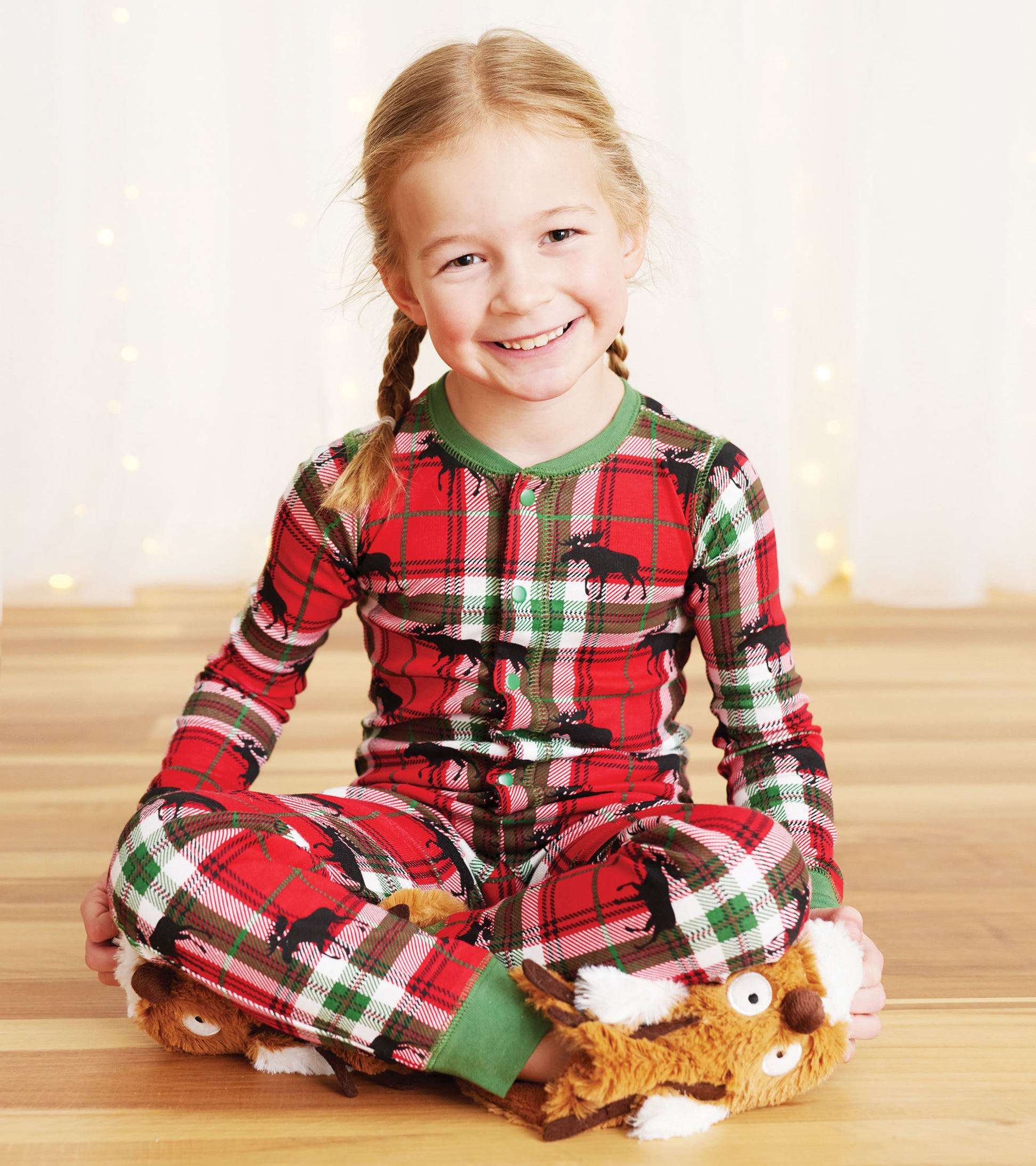 Red plaid kids' onesie featuring moose print, with green trim, worn by a smiling girl sitting cross-legged.