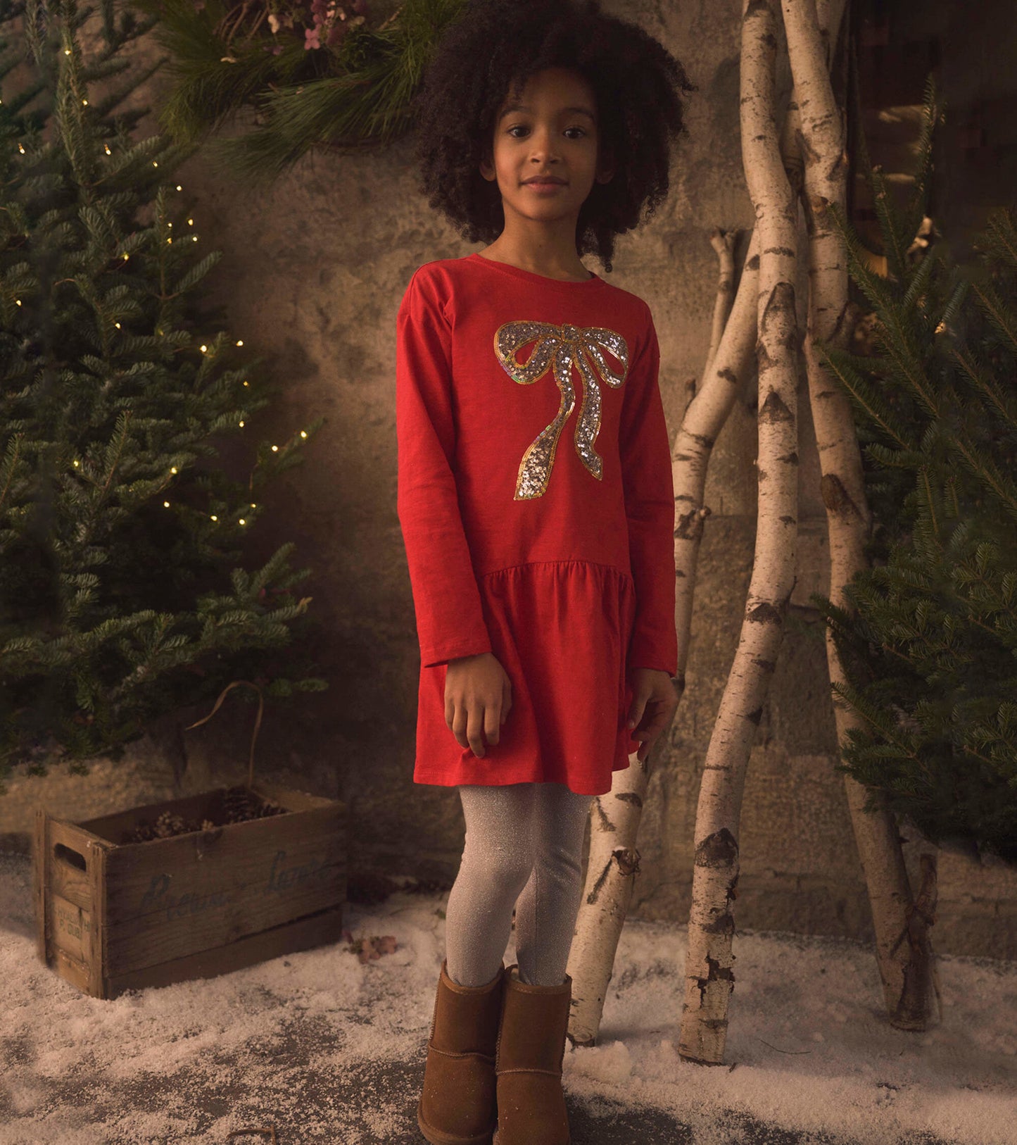 Red long-sleeve party dress with a glittery bow detail, paired with silver leggings and brown boots, set in a festive backdr…