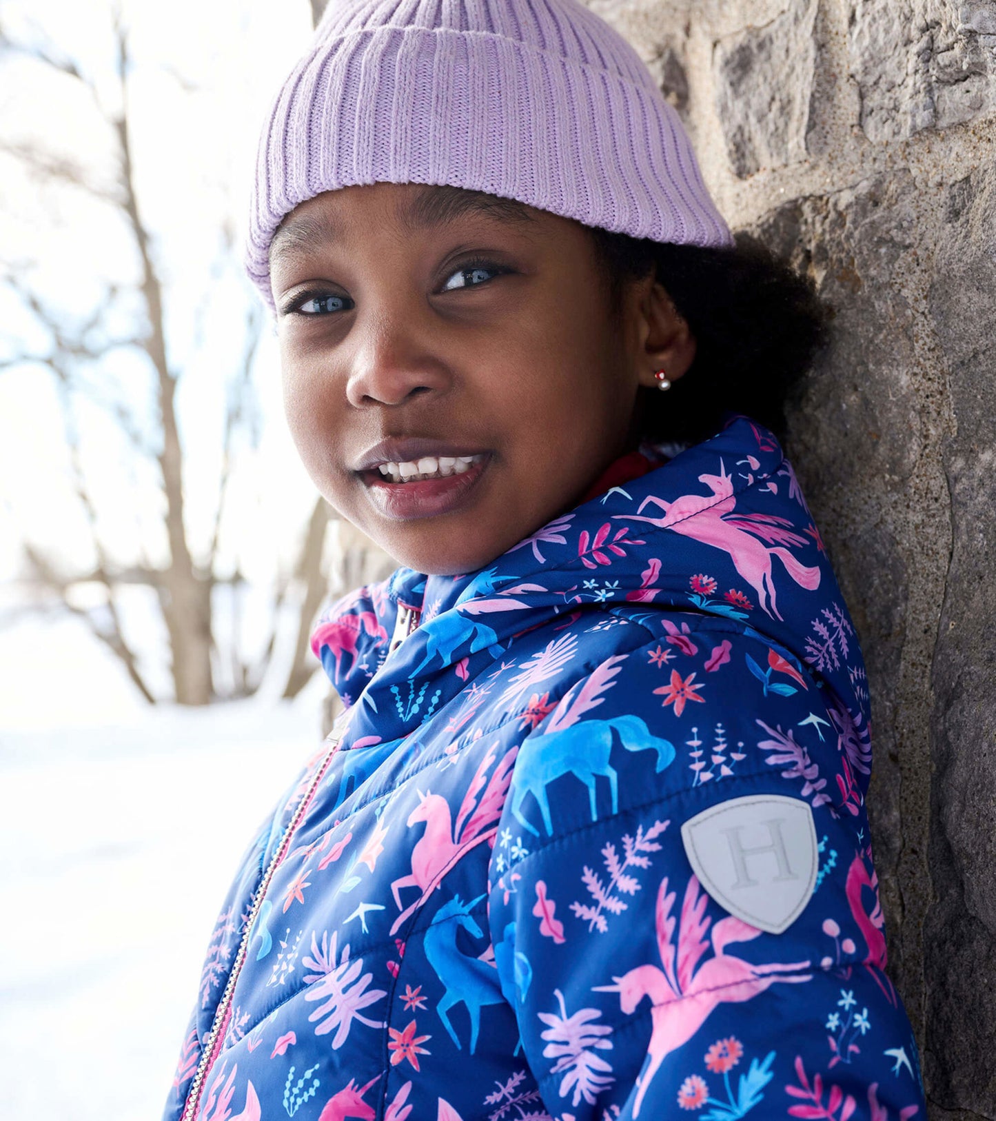 Colorful puffer jacket featuring unicorns and floral patterns, worn by a child with a lilac knit hat.