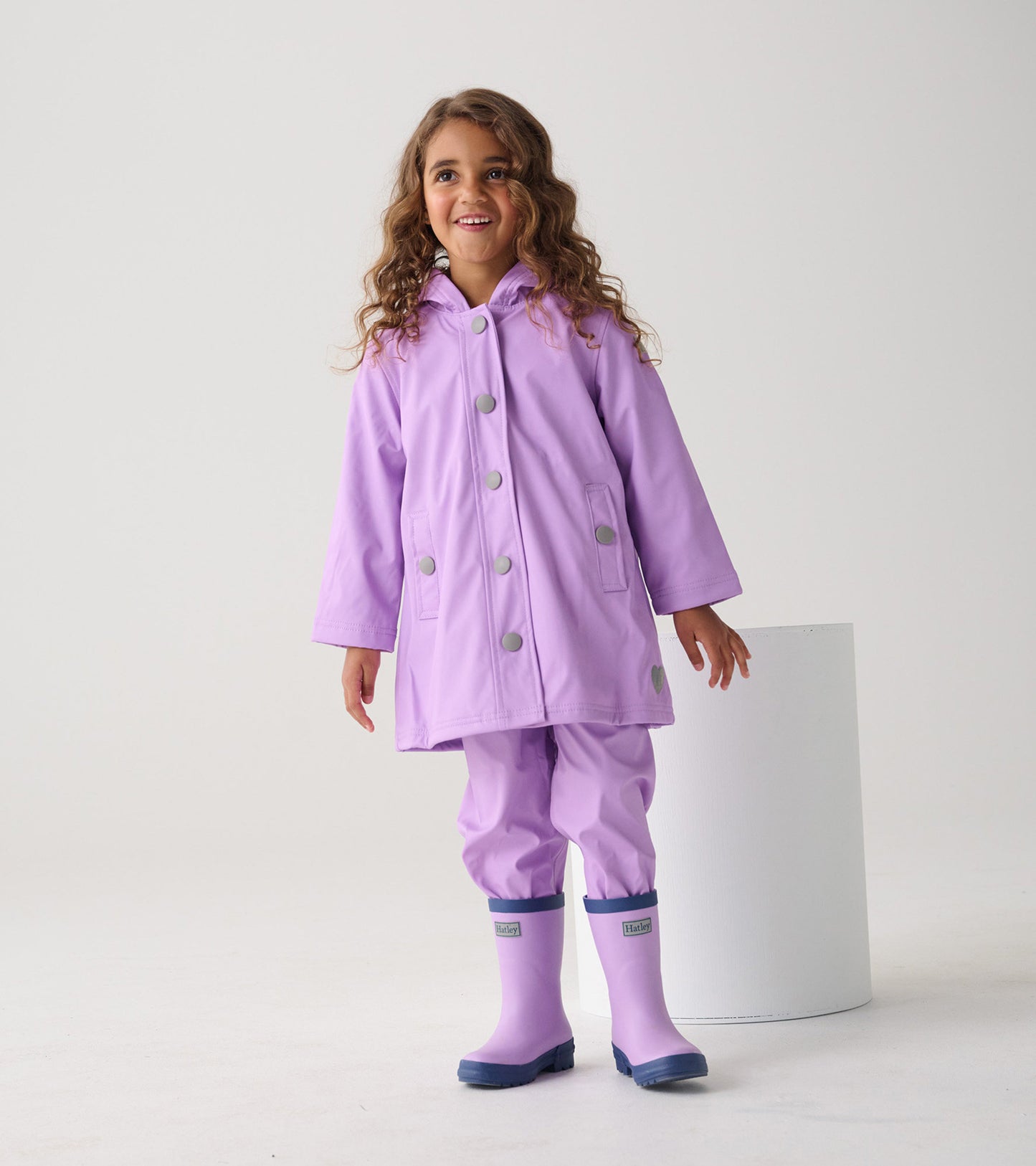 Lilac button-up rain jacket with a hood and silver buttons, worn by a smiling girl, paired with matching rain boots.