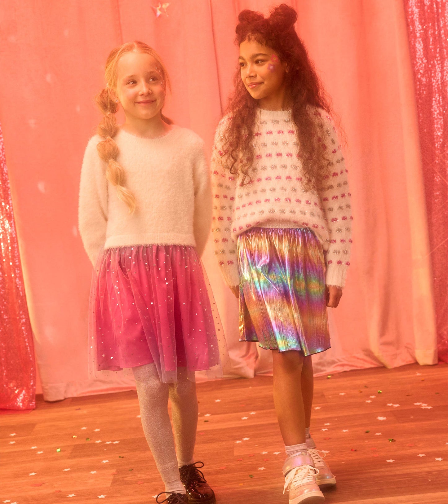 Two girls wearing fluffy sweaters, one white with colorful dots, the other with a pink tulle skirt and shiny rainbow skirt.
