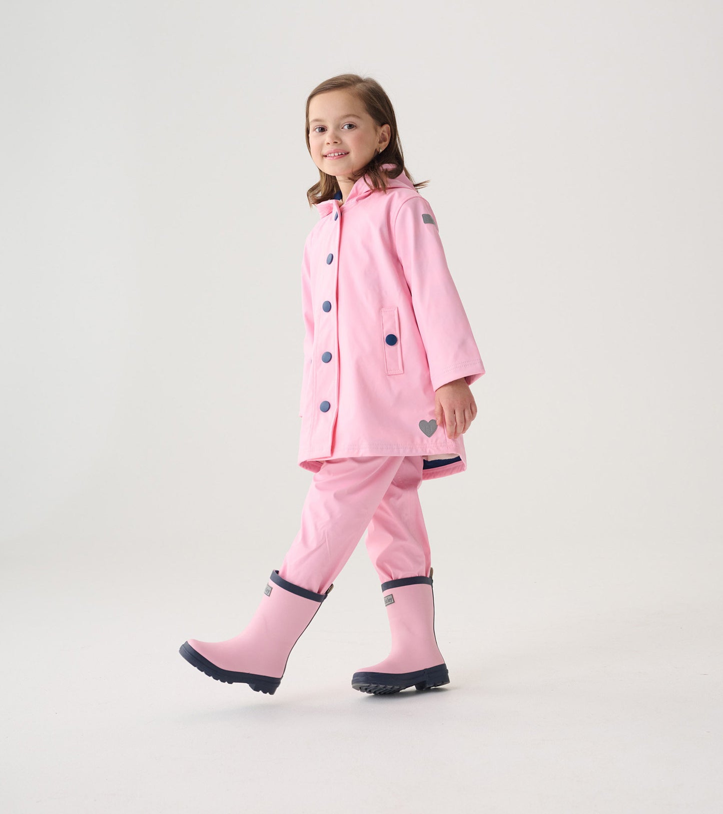 Pink button-up rain jacket with navy buttons, hood, and heart detail, worn by a girl with matching pink rain boots.