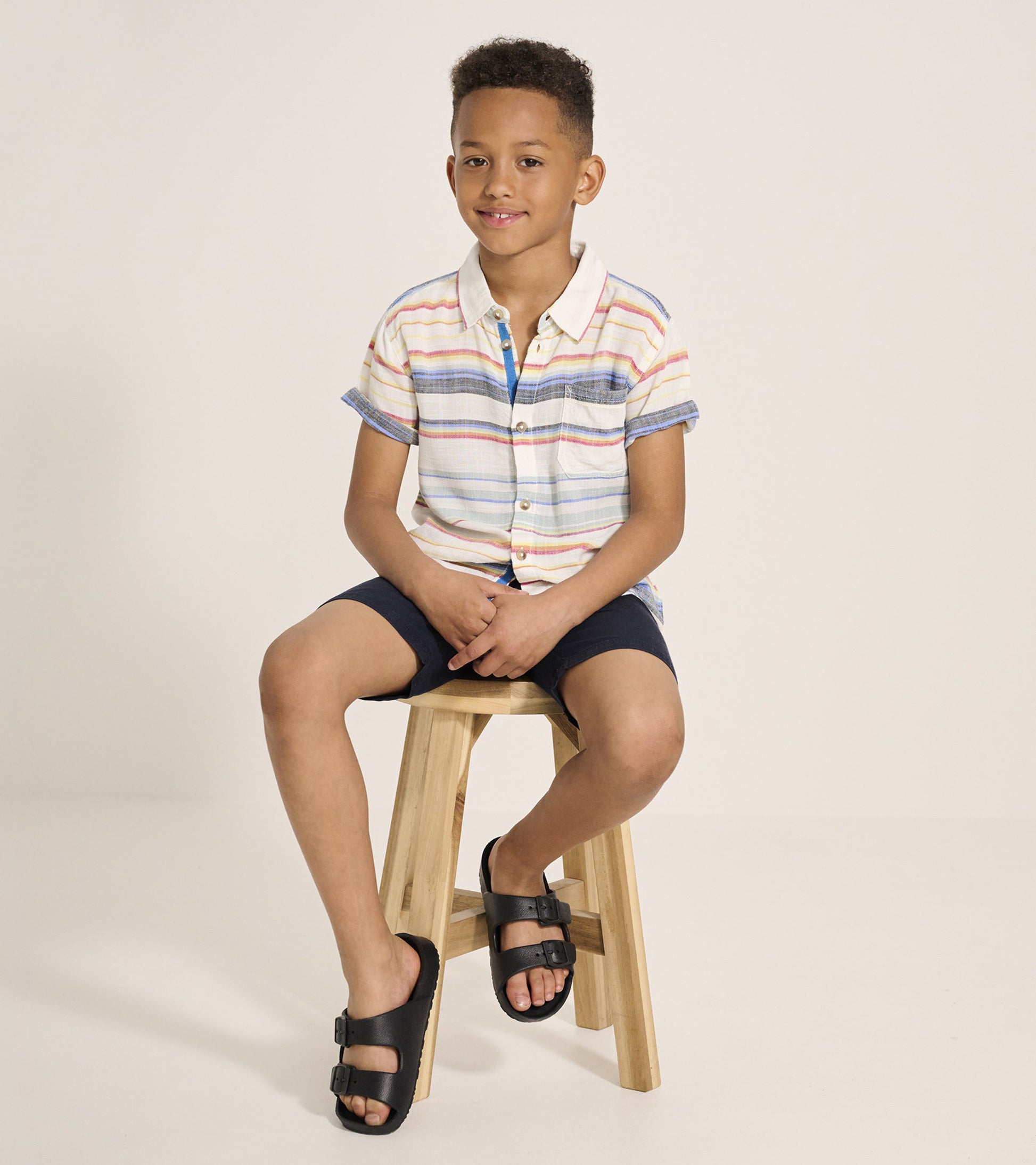 Short sleeve button-down shirt with colorful stripes, paired with navy shorts and black sandals, worn by a seated boy.