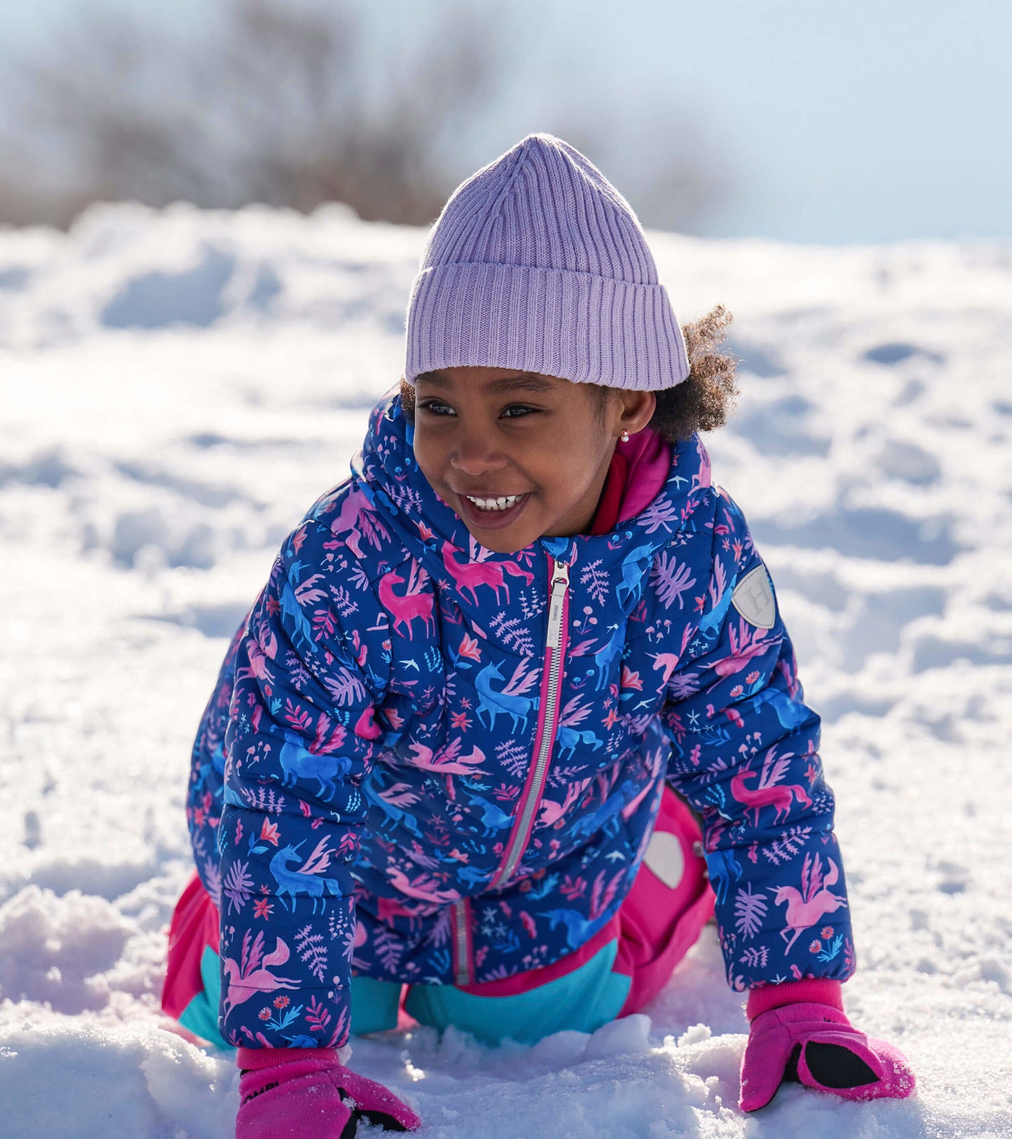Colorful puffer jacket with unicorn and floral patterns, worn by a child playing in the snow, featuring a hood and zip closu…