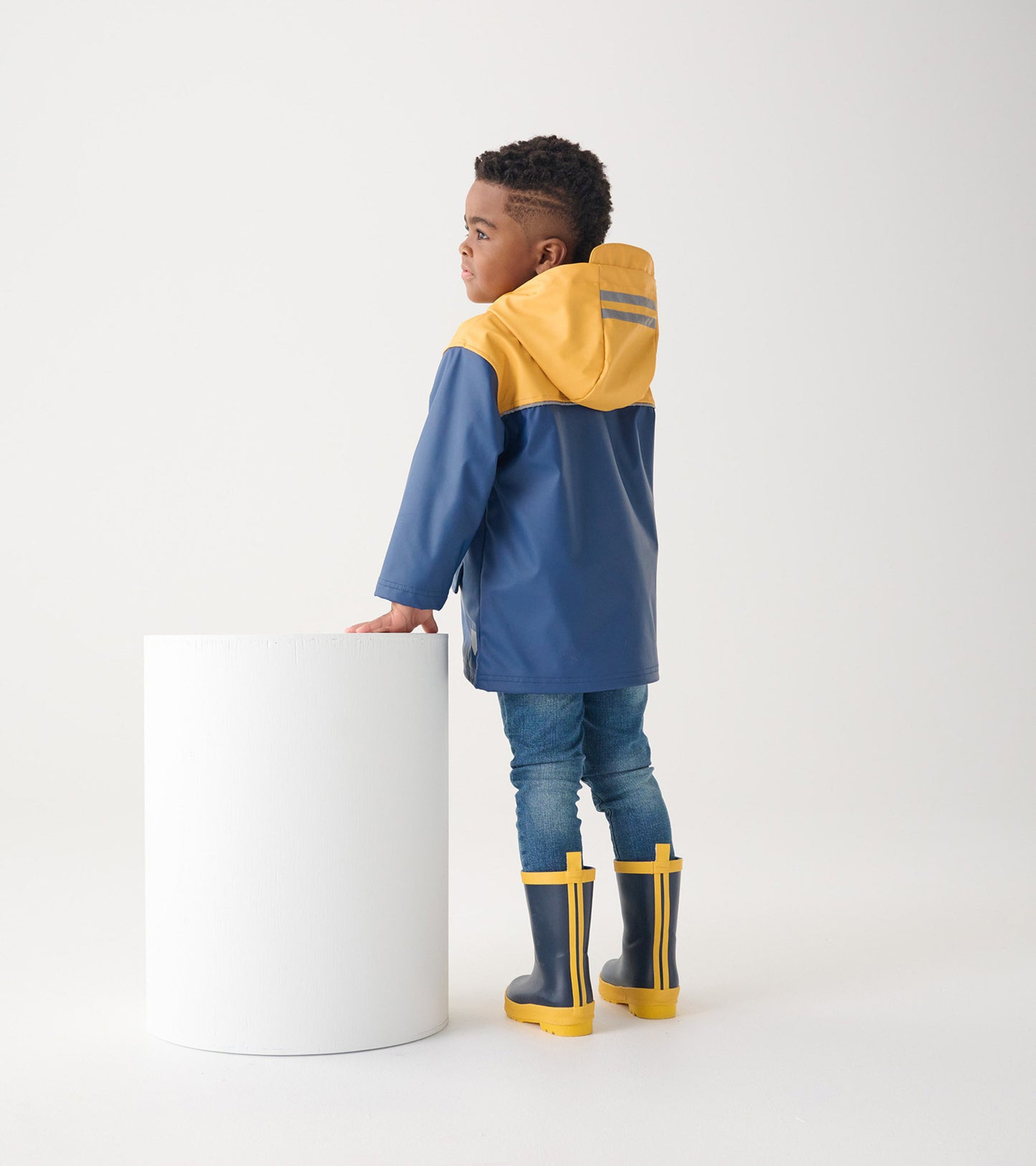 Yellow and navy zip-up rain jacket with a hood, worn by a child, paired with blue jeans and yellow rain boots.