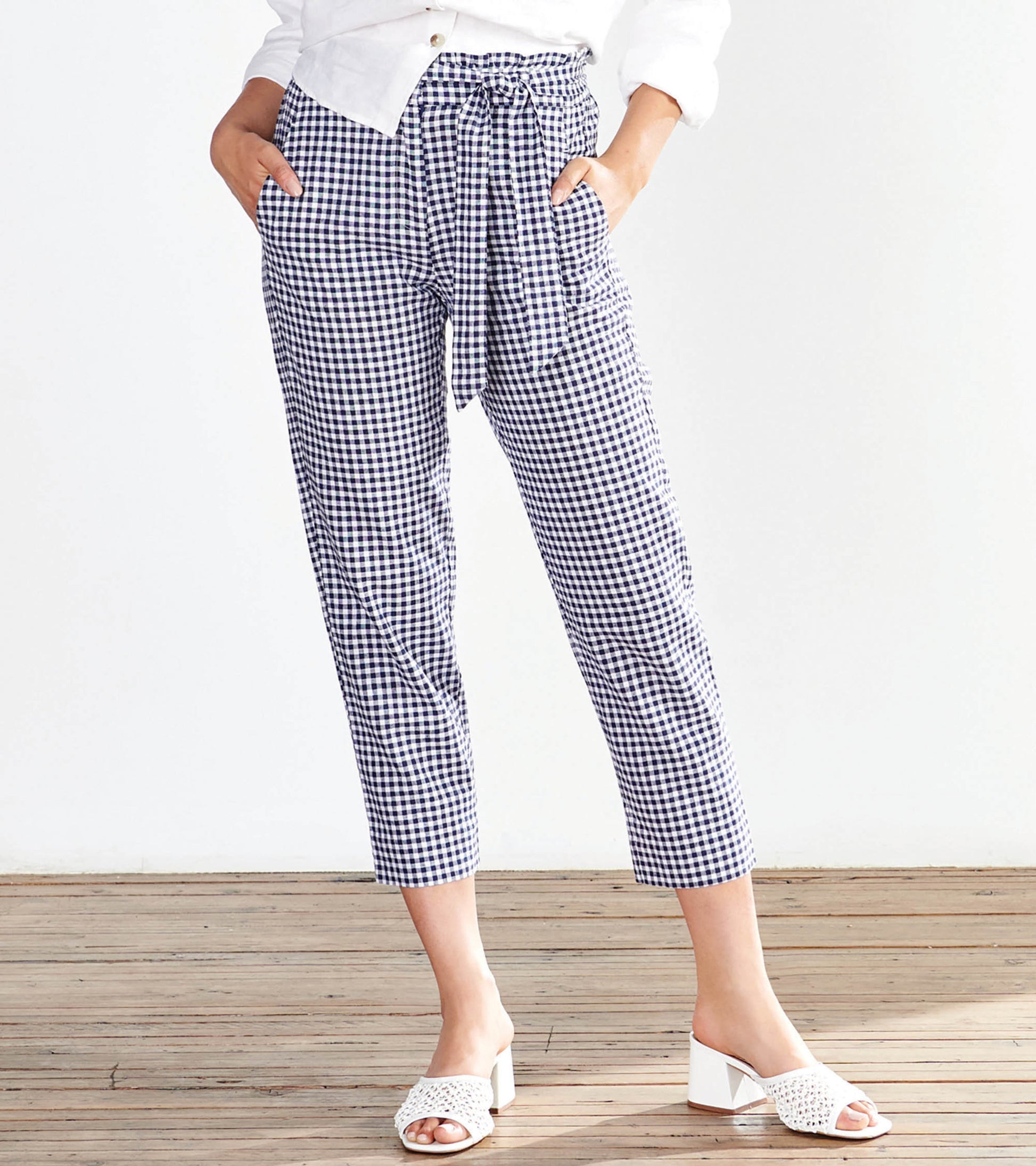 Navy gingham paper bag pants with a tie waist and side pockets, styled with a white top and heeled sandals.