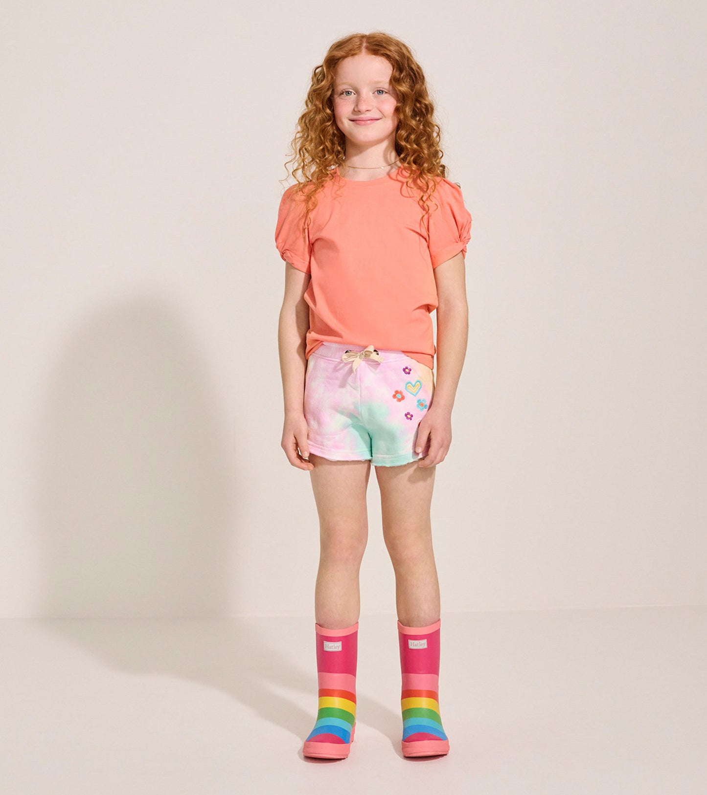 Colorful tie-dye shorts with floral and heart designs, paired with a coral shirt and rainbow rain boots.