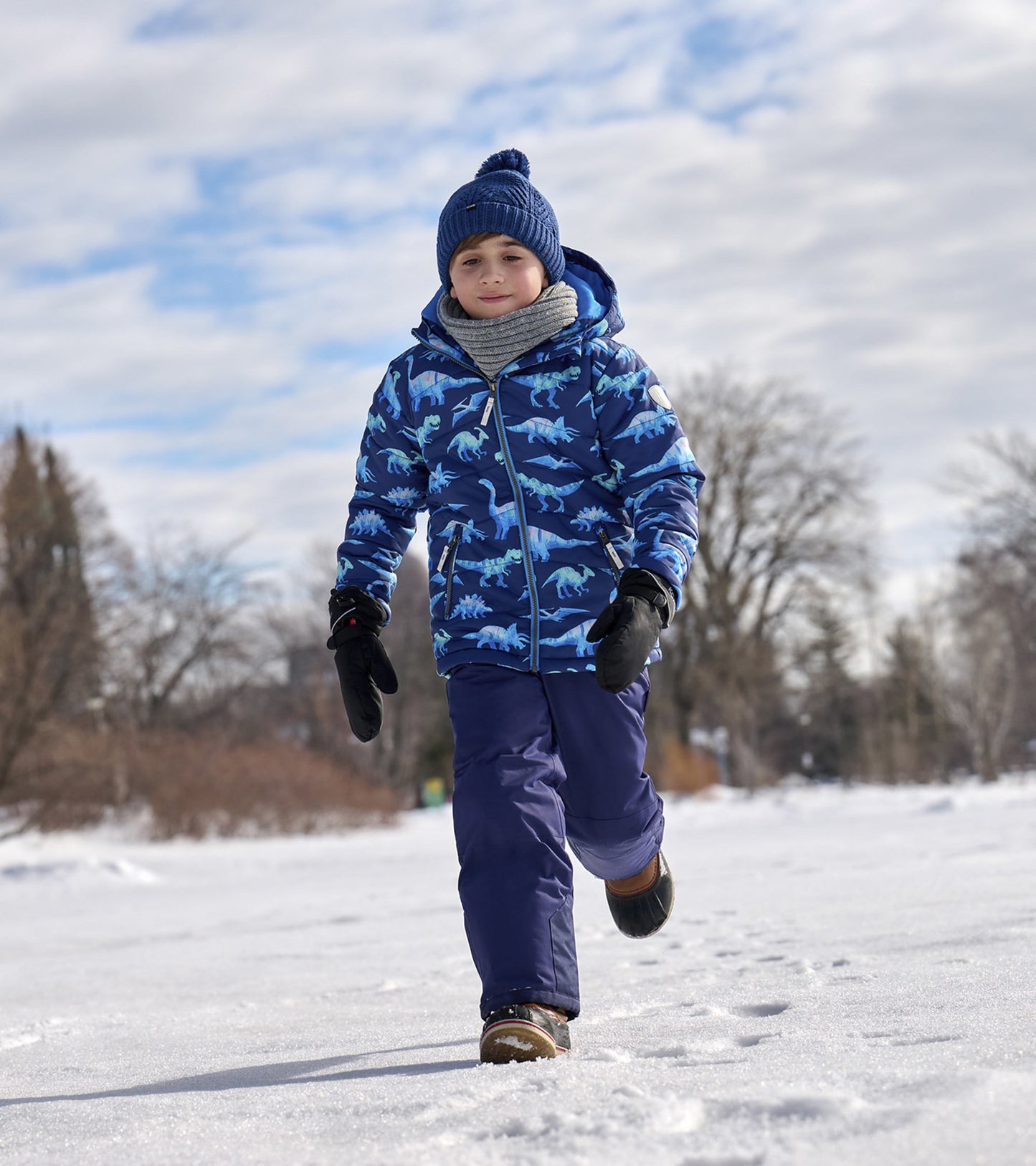 Blue dinosaur-patterned puffer jacket worn by a child, paired with gloves and a knitted hat, walking in the snow.