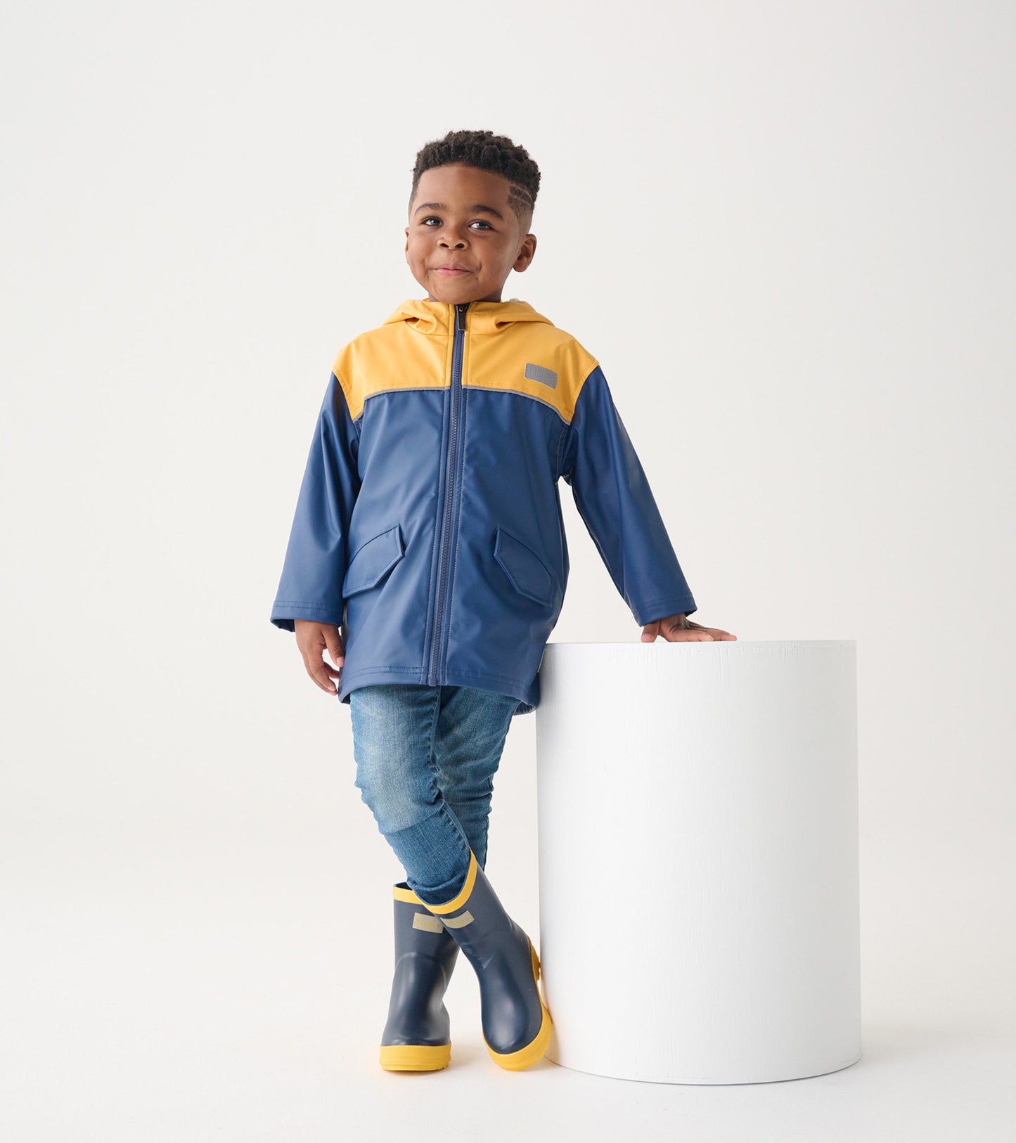 Yellow and navy zip-up rain jacket on a child, featuring a hood and front pockets, paired with blue jeans and rain boots.
