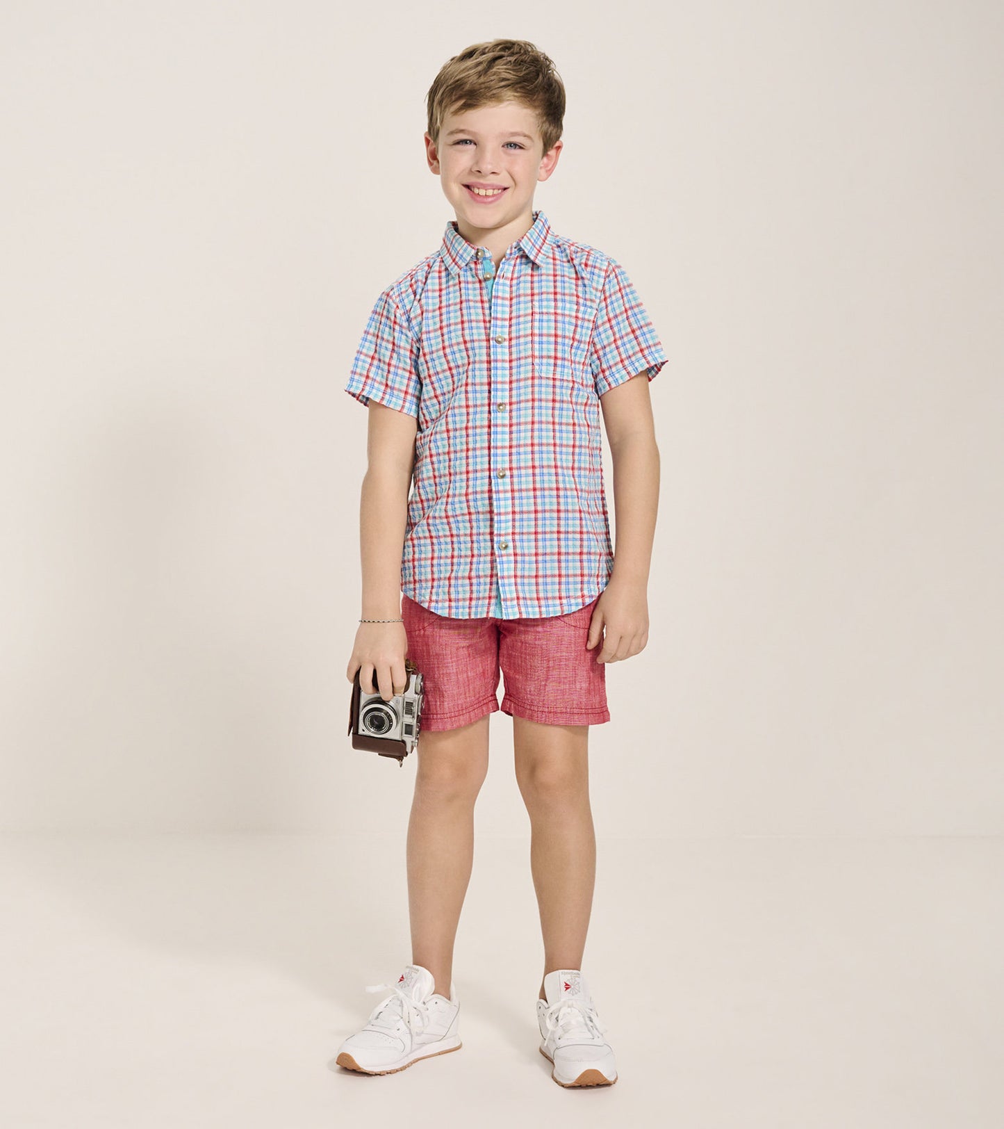 Short-sleeve button-down shirt featuring a blue, red, and white check pattern, worn with red shorts and white sneakers.