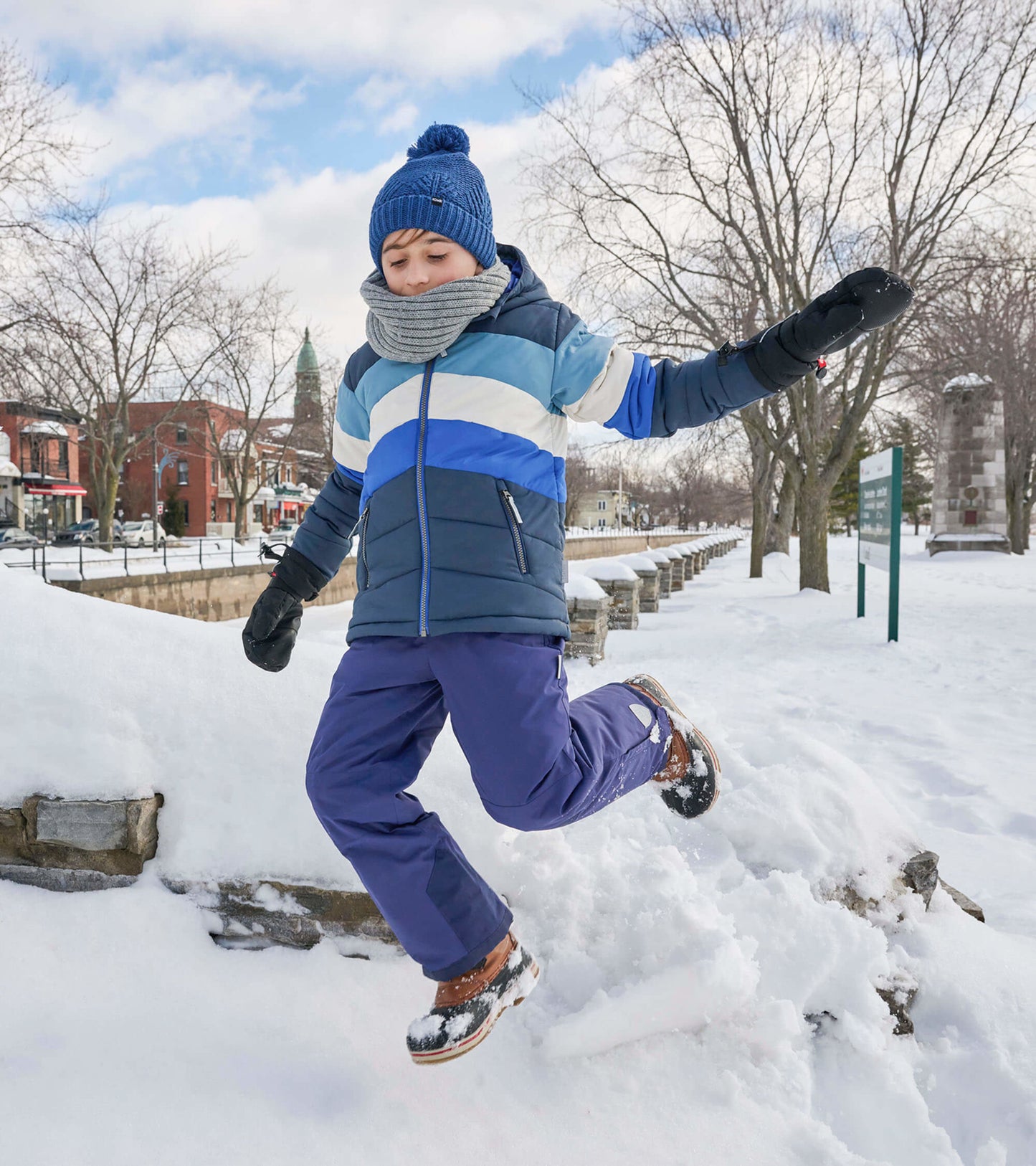 Black and blue puffer jacket with horizontal stripes, worn by a child jumping in snow, paired with gloves and a hat.