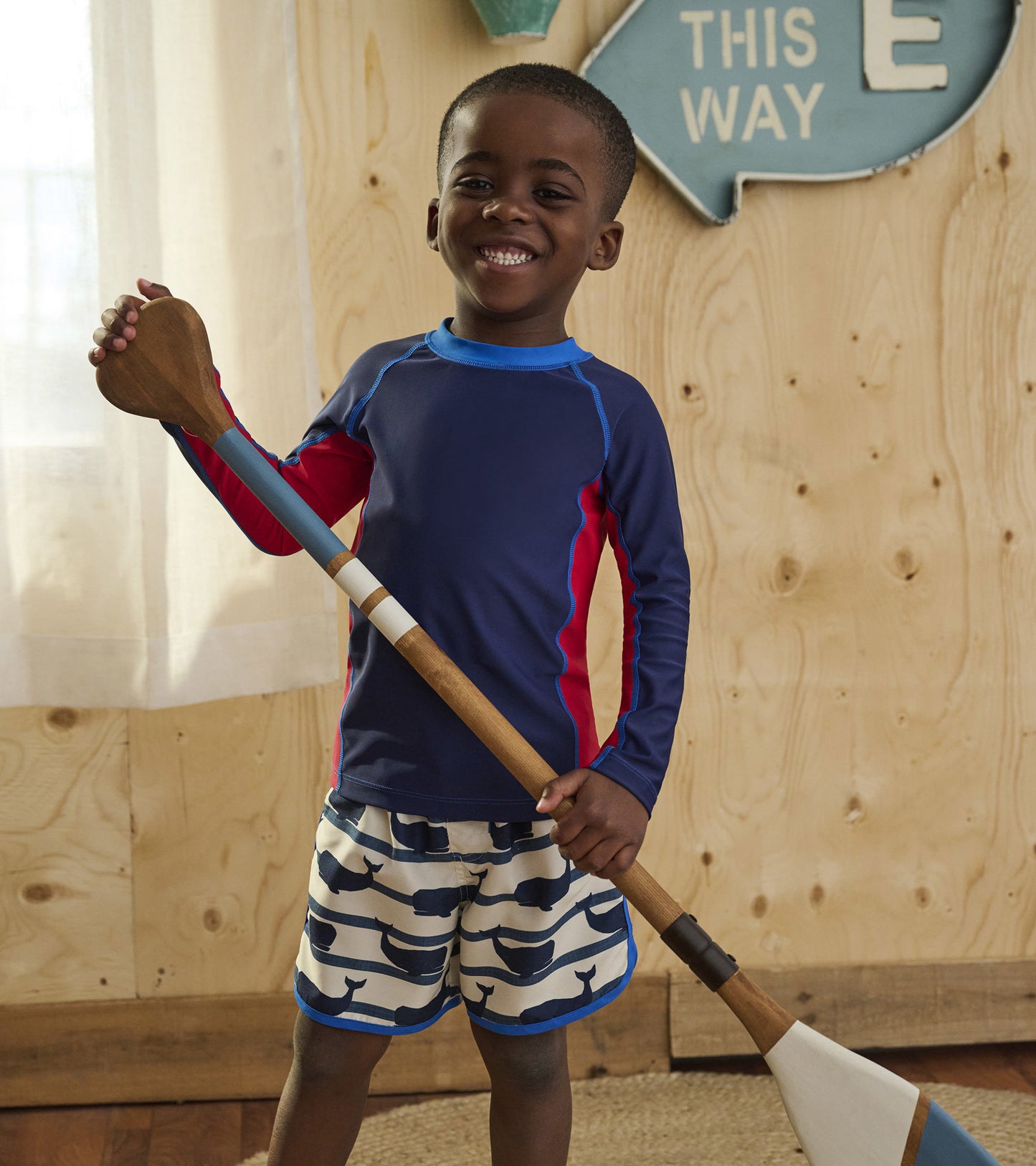Navy long sleeve rashguard with red accents, paired with whale-patterned shorts, worn by a smiling boy holding a paddle.