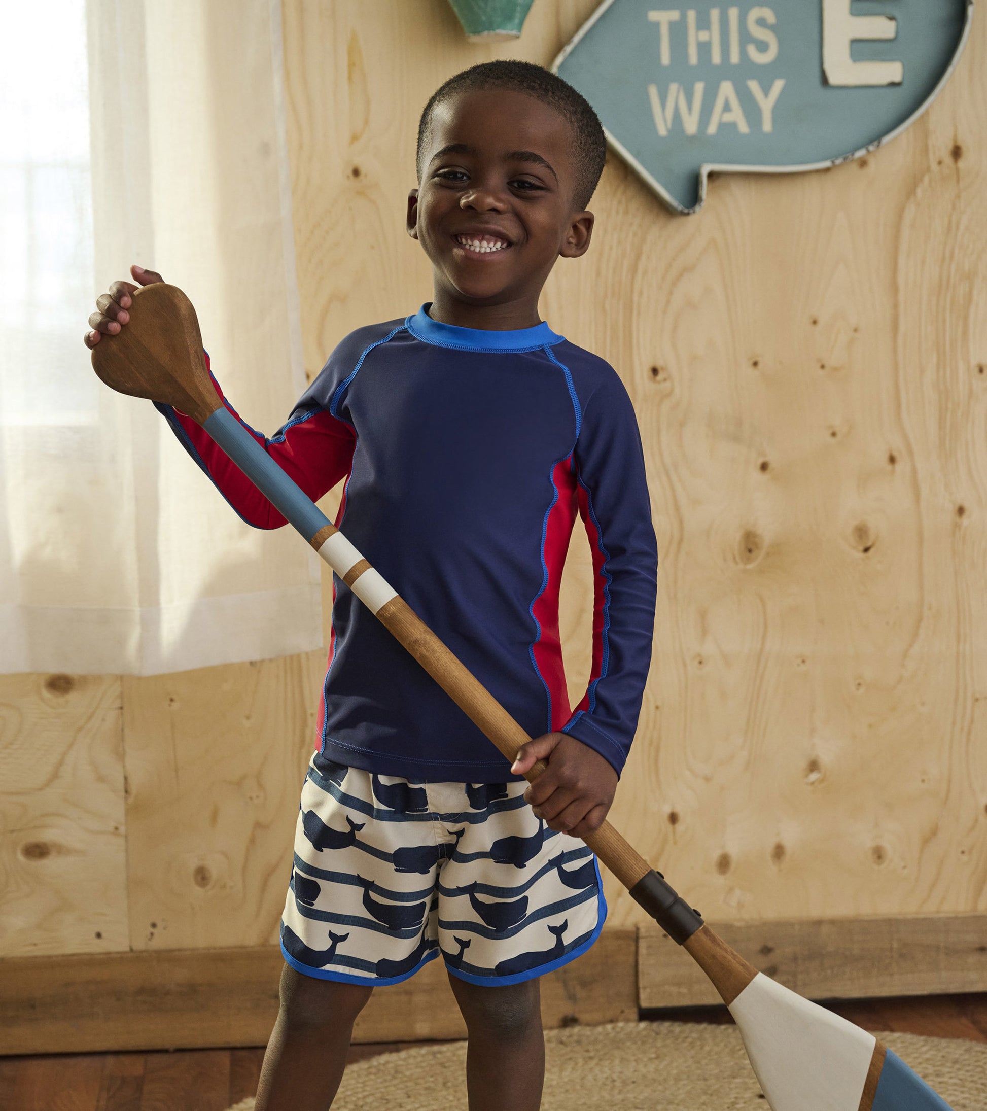 Navy long sleeve rashguard with red accents, paired with whale-patterned shorts, worn by a smiling boy holding a paddle.