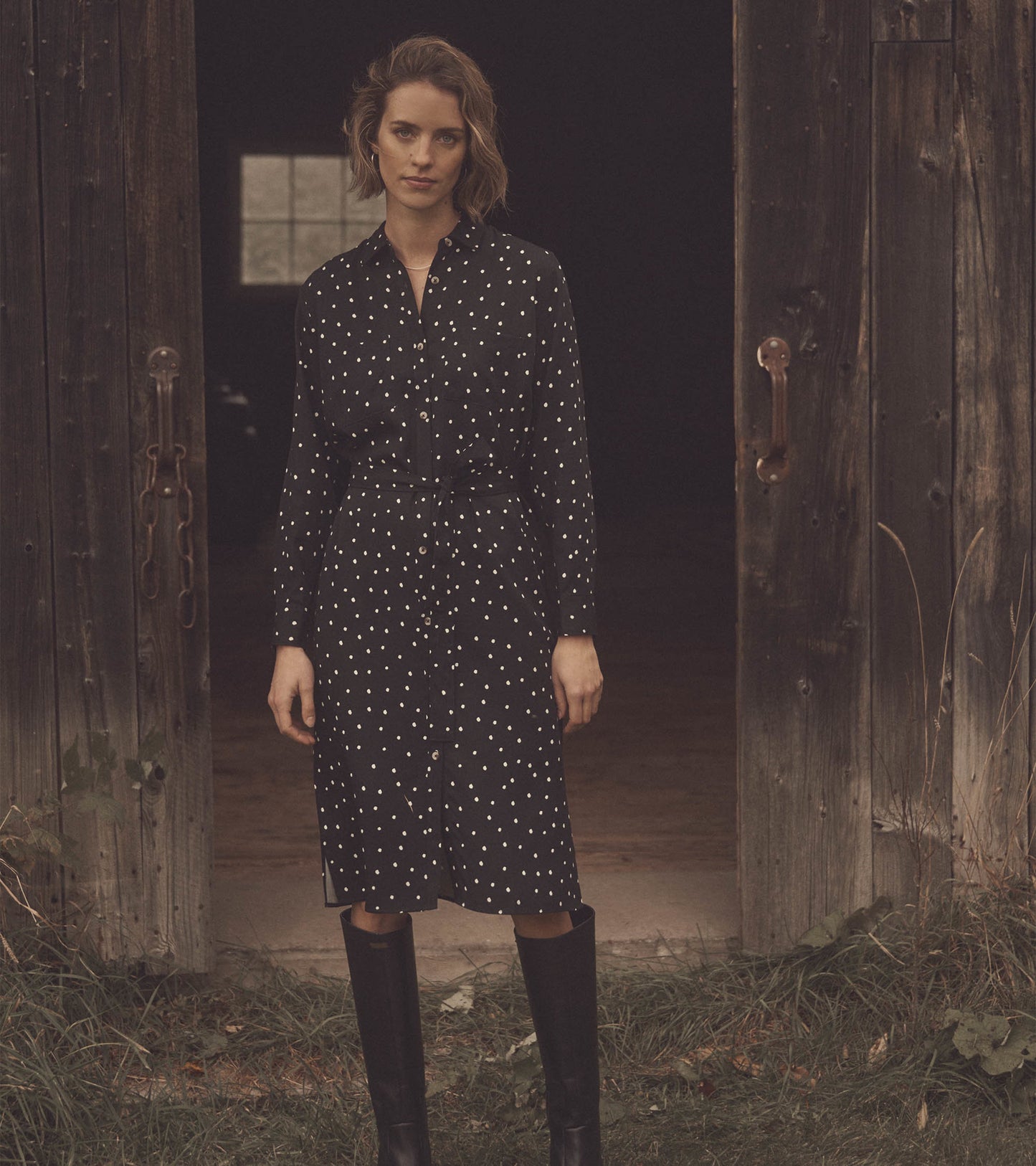 Black midi shirt dress with white spots, featuring a button-down front and belted waist, styled with knee-high boots.