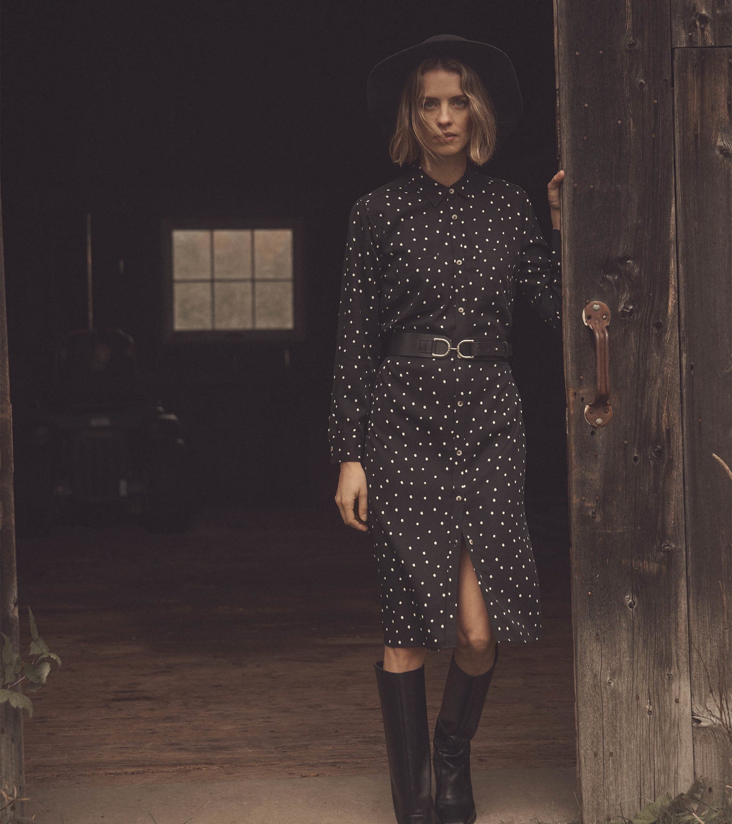 Black midi shirt dress with white spots, featuring a button front, belted waist, and a side slit. Styled with a hat and boot…